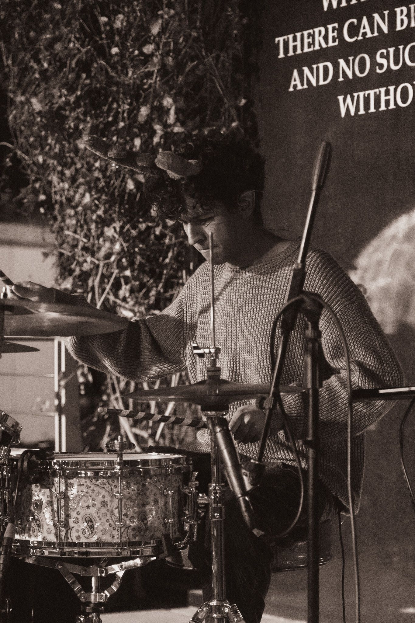 A drummer plays behind a drum kit in black and white, performing on stage with text visible on a wall behind them.