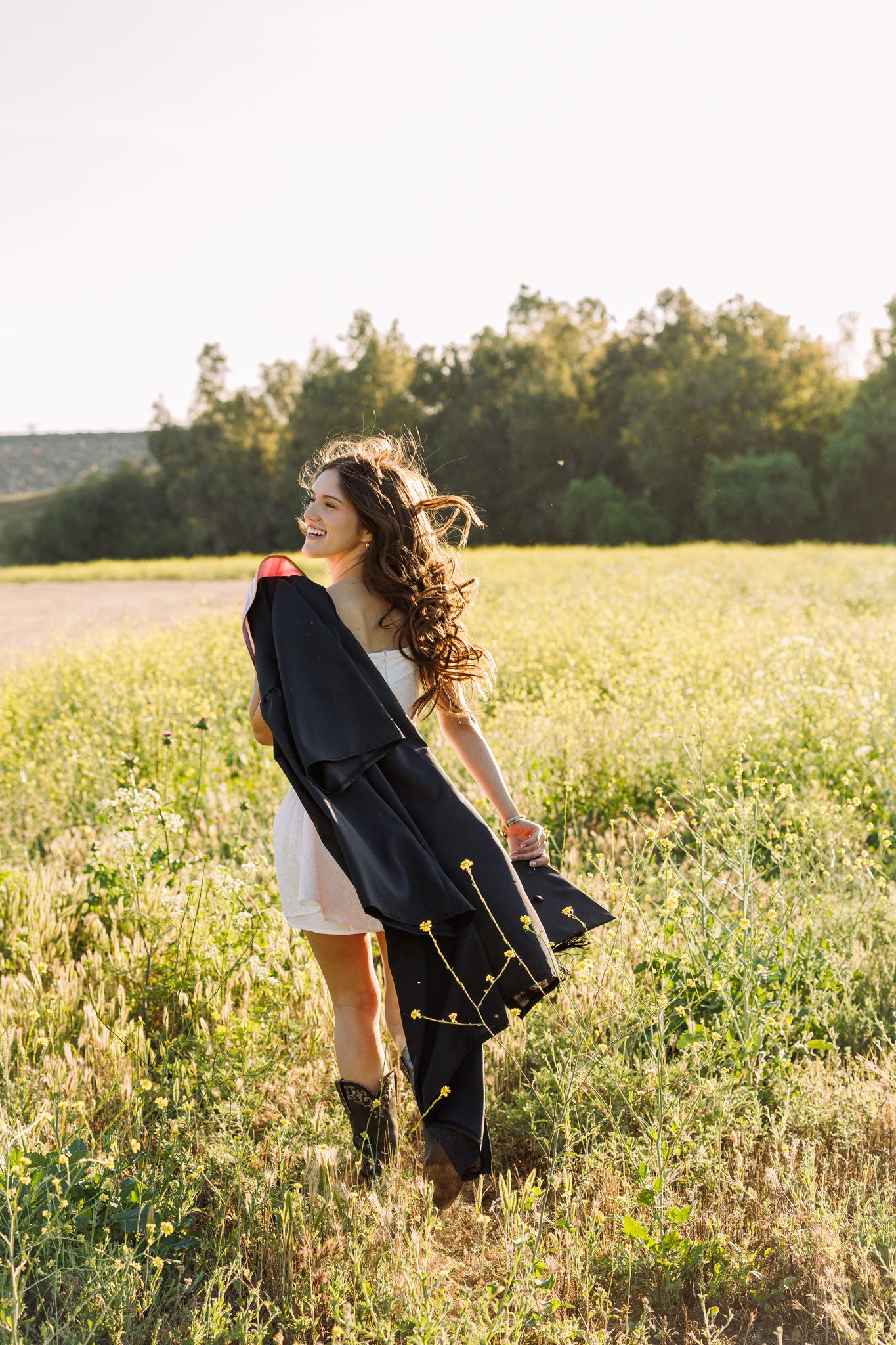 Smiling young woman in white dress and graduation gown standing in sunny green field with curly hair blowing in the wind.