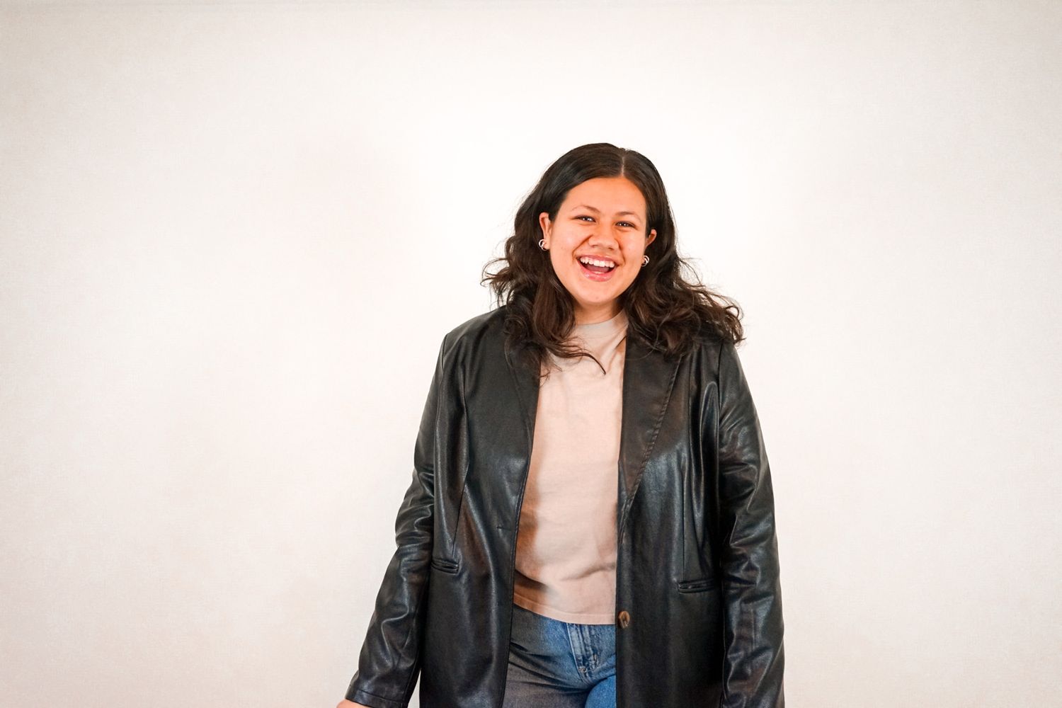 A person wearing a black leather jacket and light top smiles warmly against a plain white background.