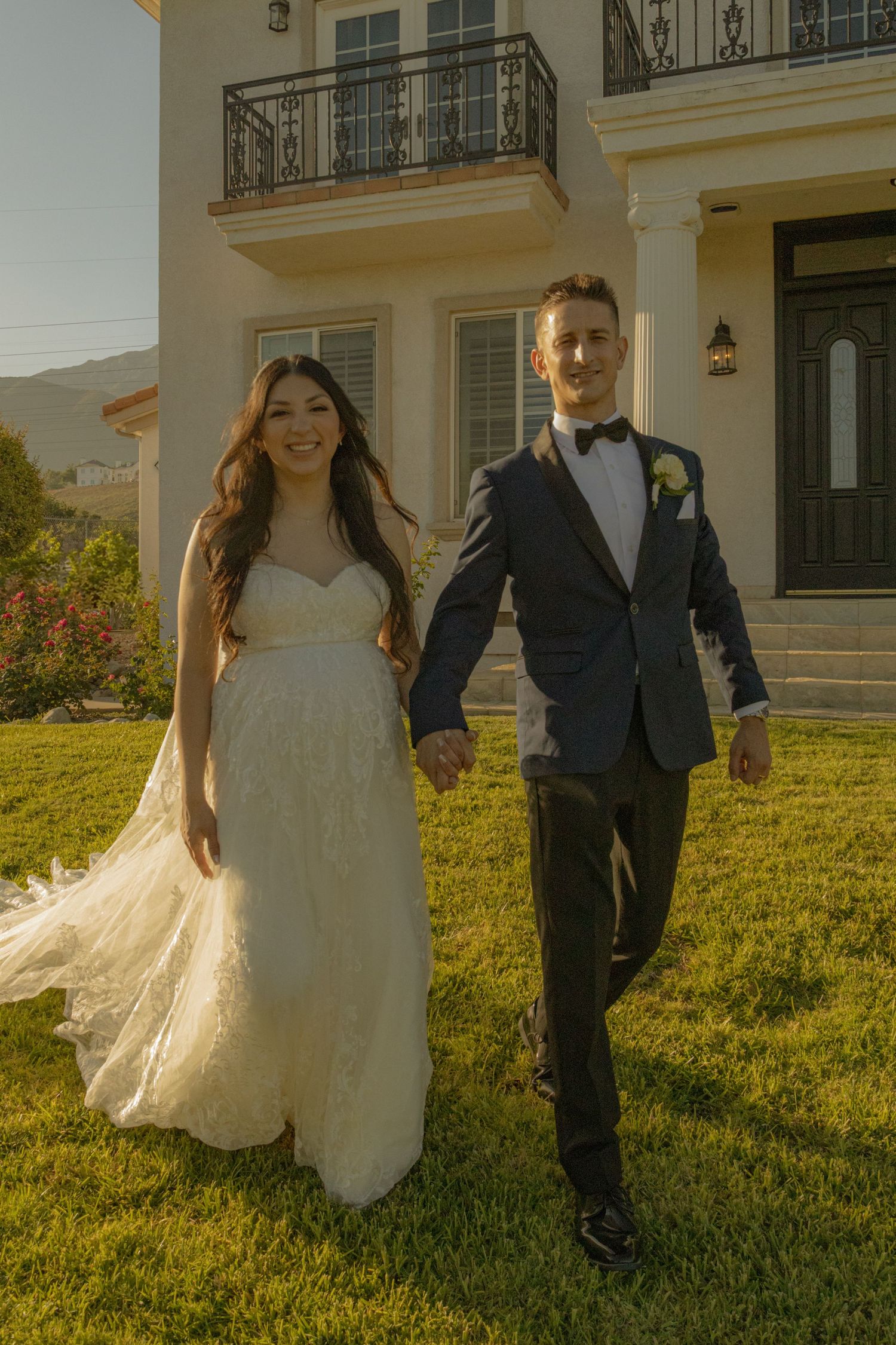 A bride in a flowing white dress and groom in a dark tuxedo walk hand-in-hand on a sunny lawn outside an elegant home.