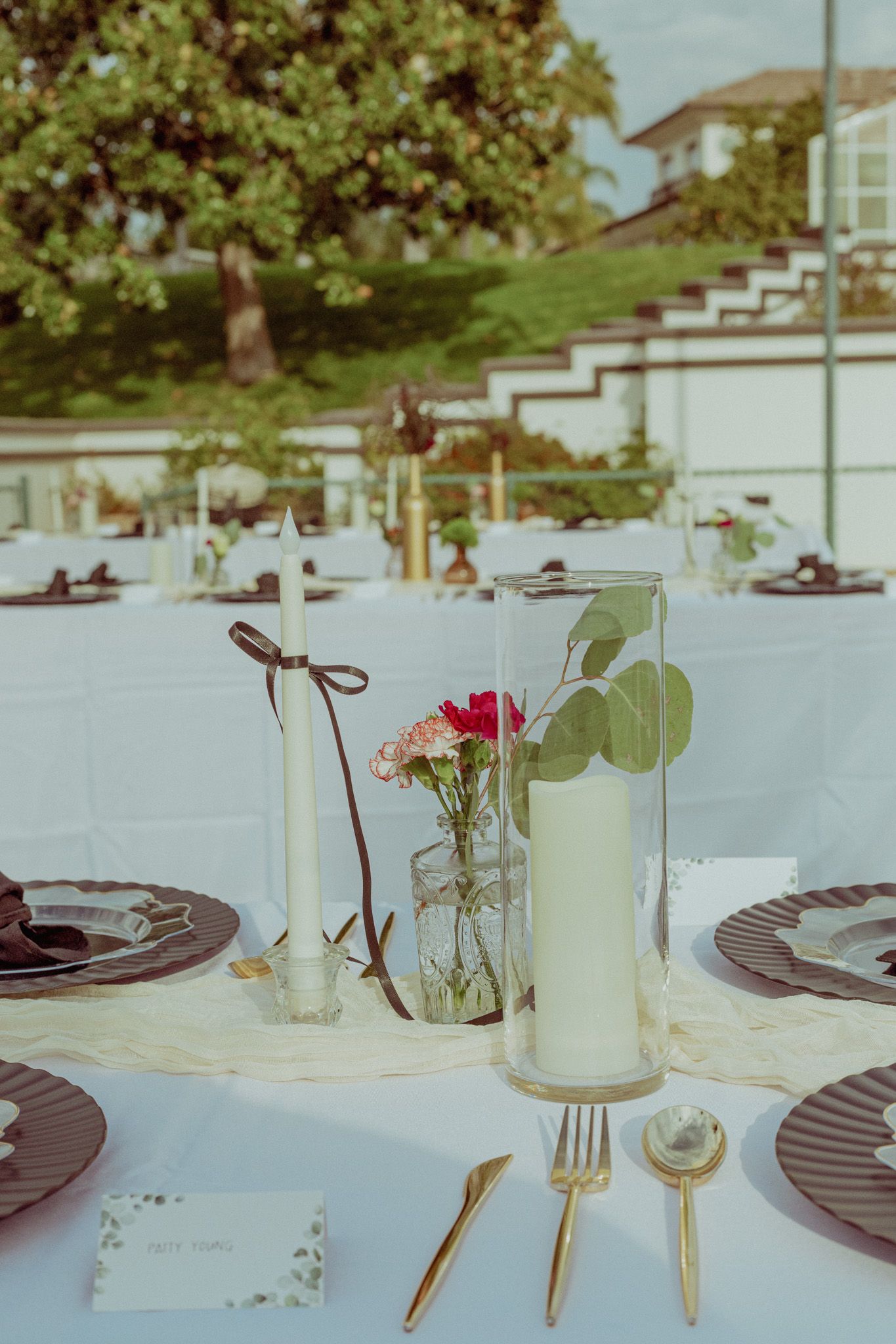 Elegant outdoor table setting with white linens, glass vases with flowers, and black plates on a sunny day.