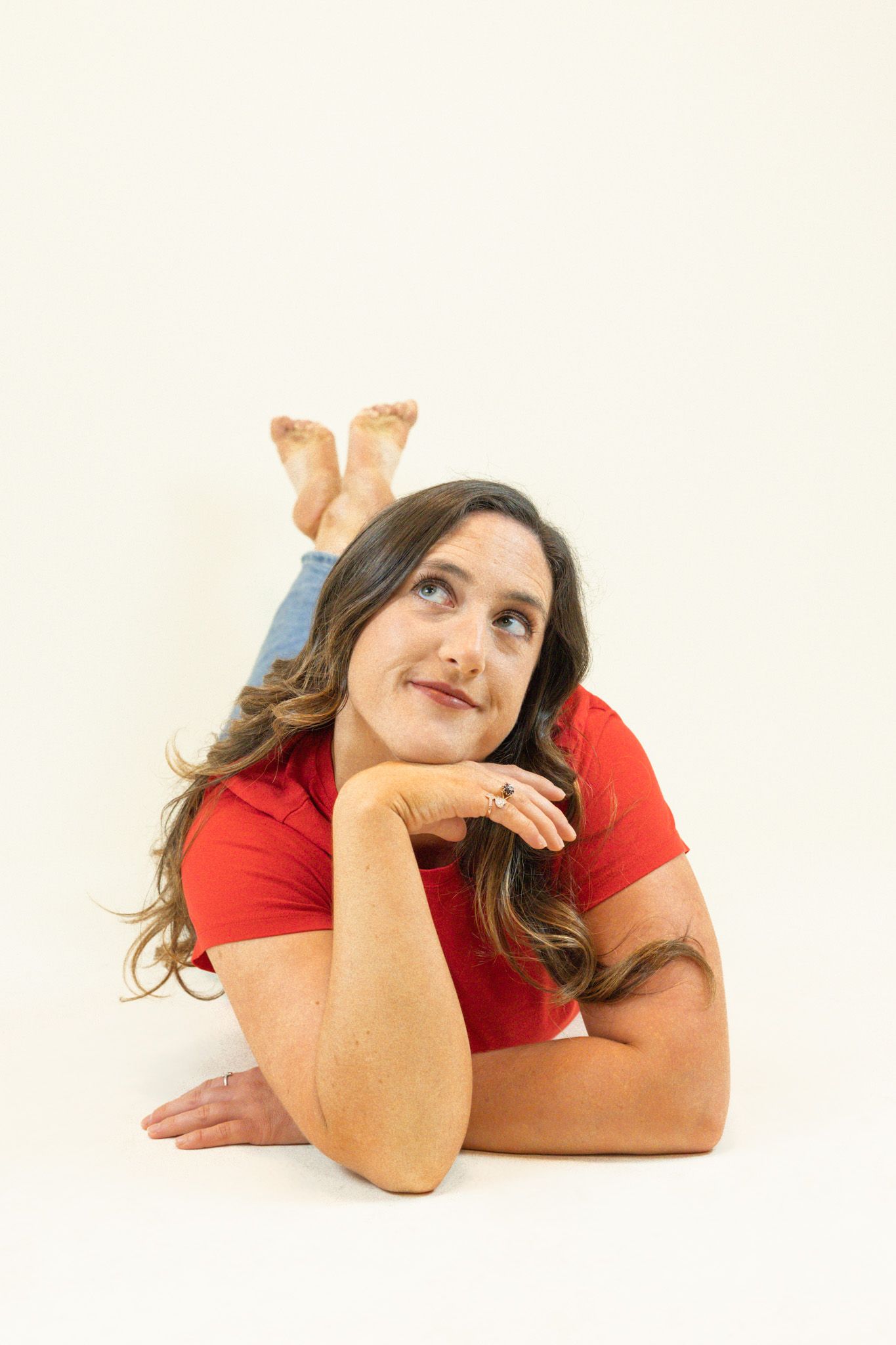 A person in a red shirt lies on a white background with feet up, looking thoughtfully away with a dreamy expression.