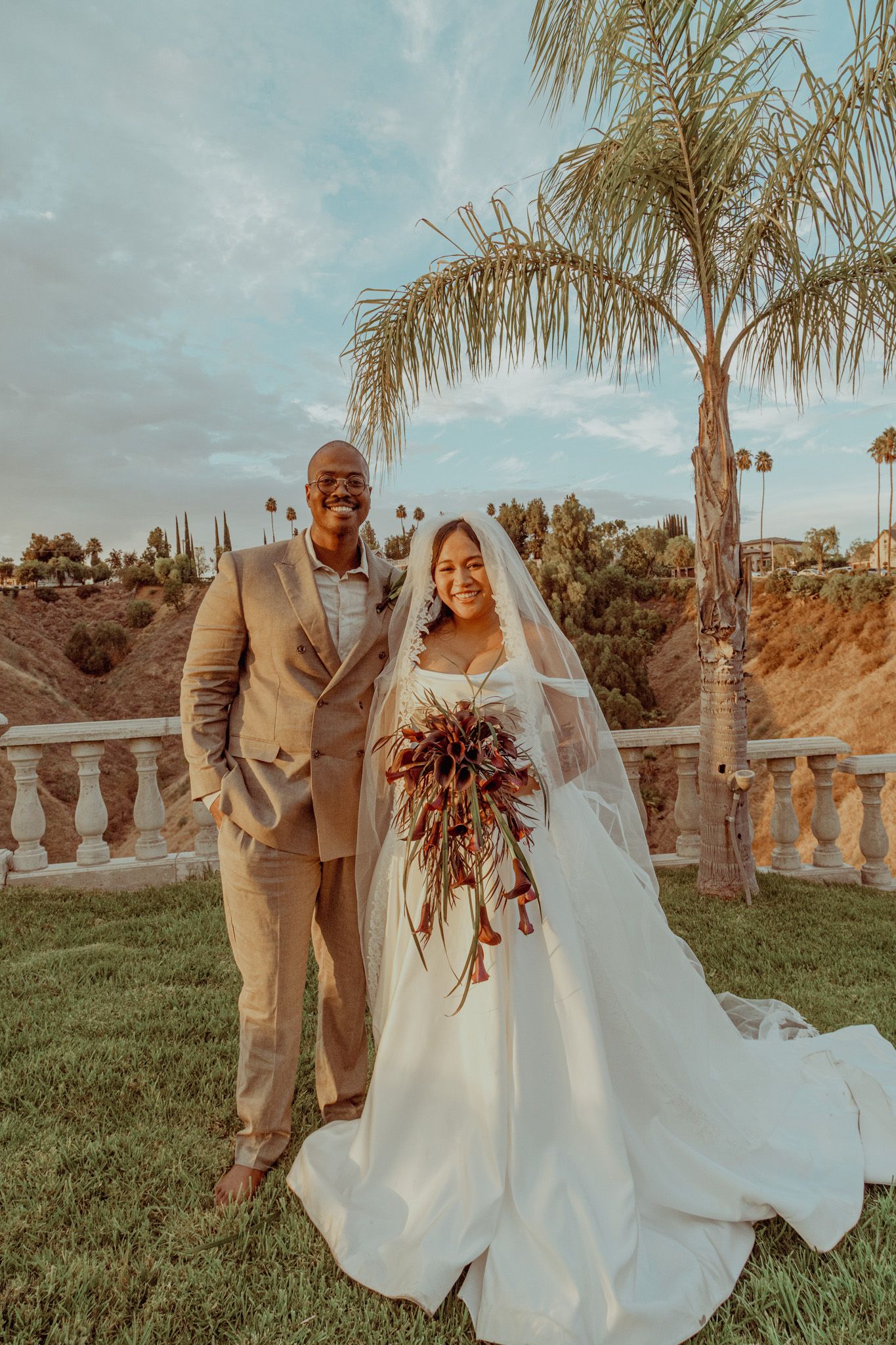 A newlywed couple poses outdoors at sunset with palm trees and rolling hills in the background.