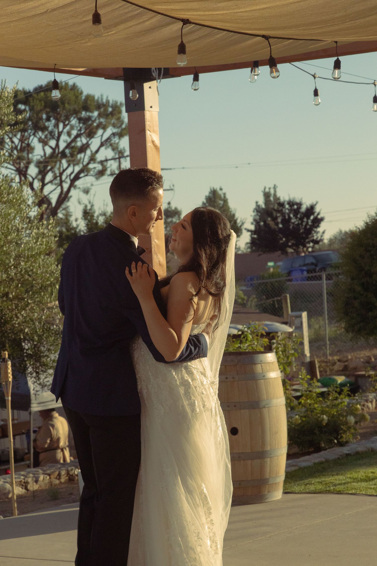 A couple shares their first dance under string lights at an outdoor wedding venue with wine barrels in the background at sunset.