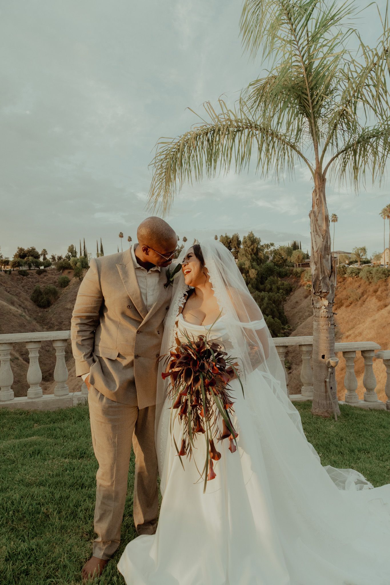 A romantic outdoor wedding scene with a couple embracing among palm trees at sunset.