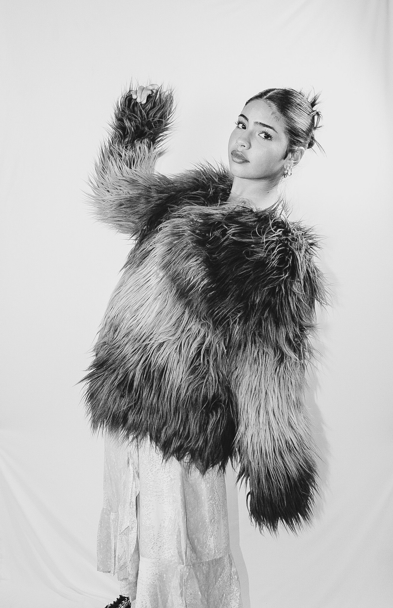 A black and white fashion photo of someone posing in a dramatic fluffy faux fur coat over a light dress.