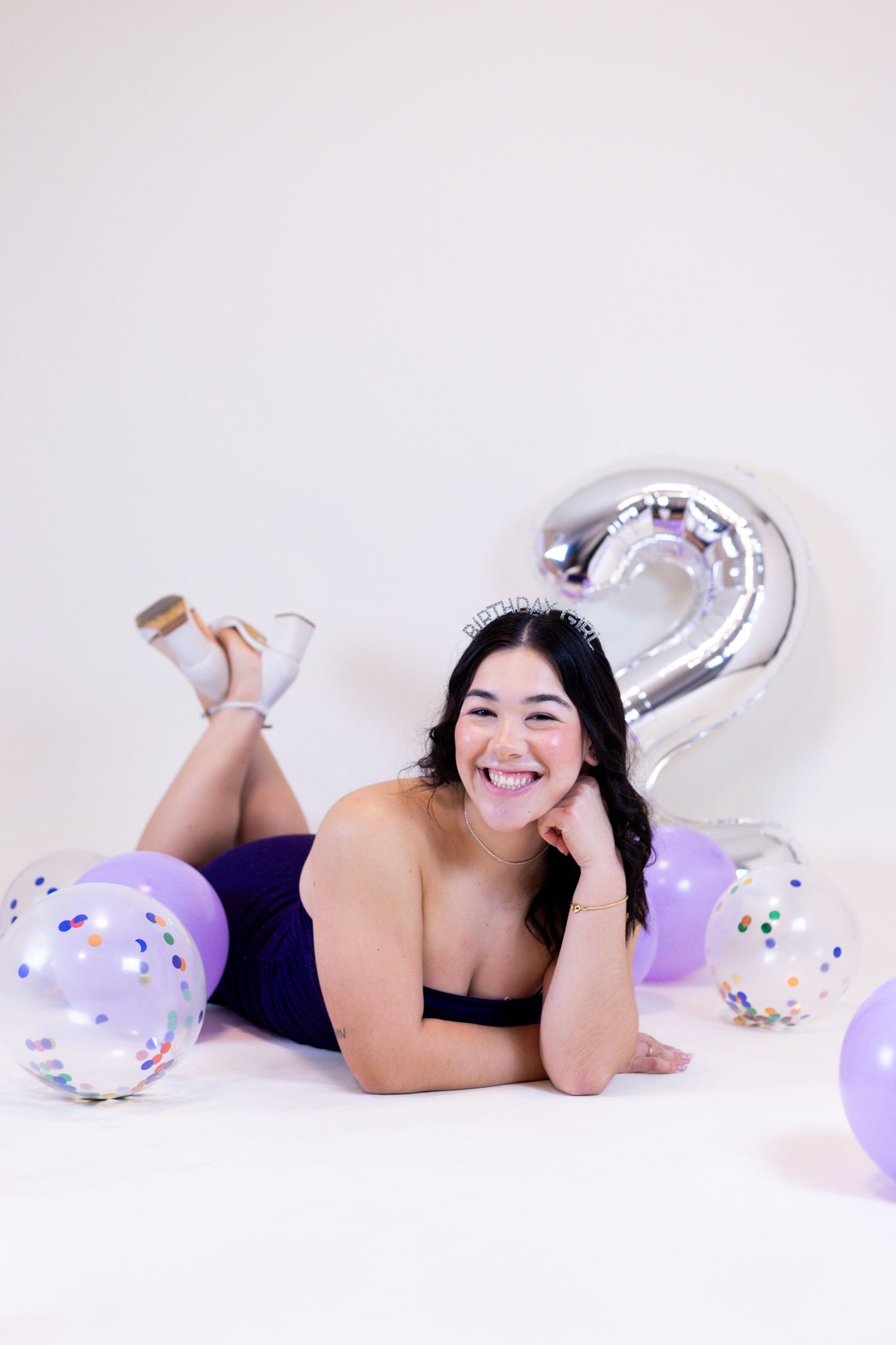 A person in black loungewear poses playfully on a white backdrop with purple balloons and confetti decorations.