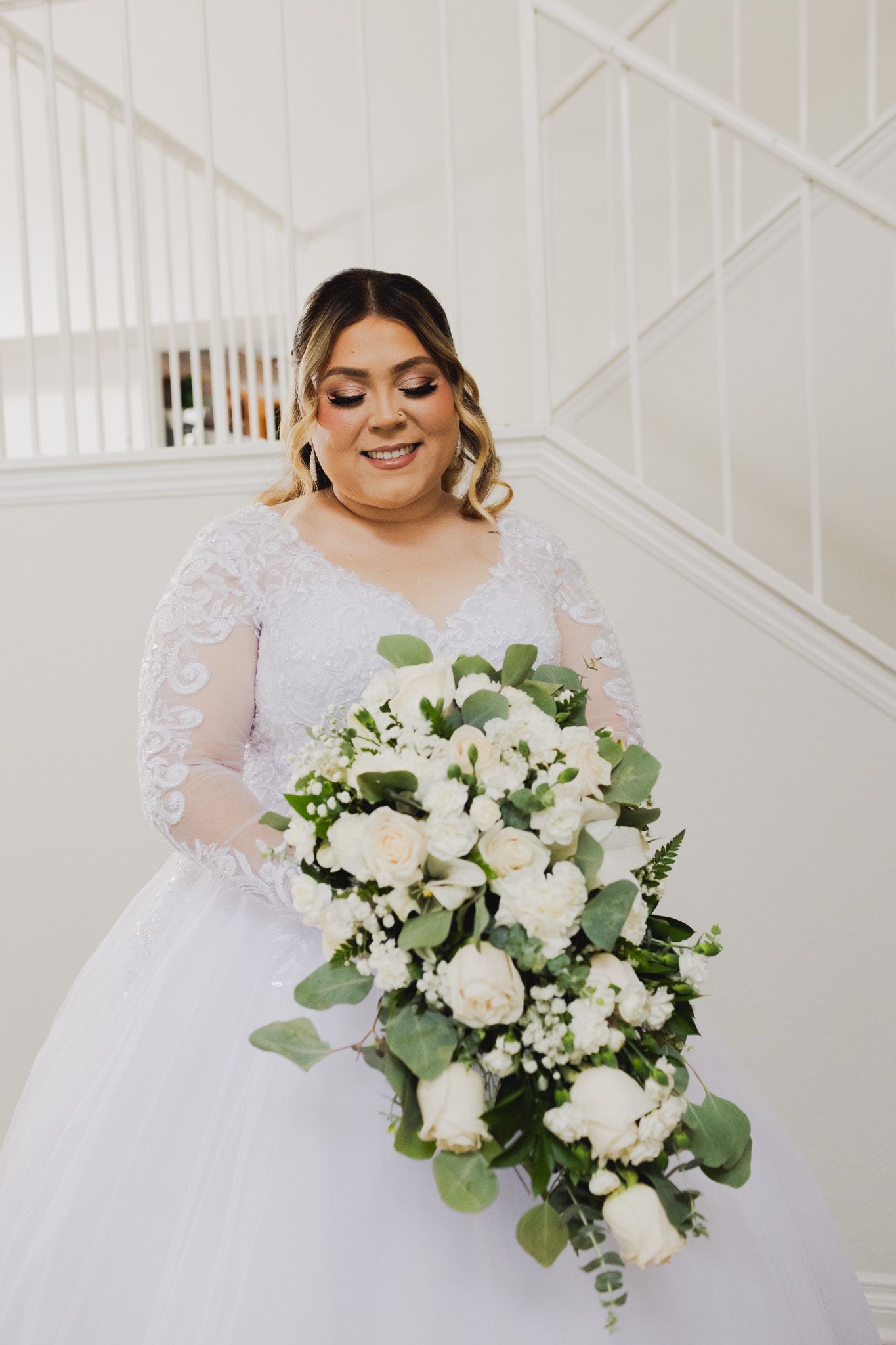 A lovely bride in a white lace gown holds a cascading bouquet of white roses, eucalyptus leaves and baby's breath.