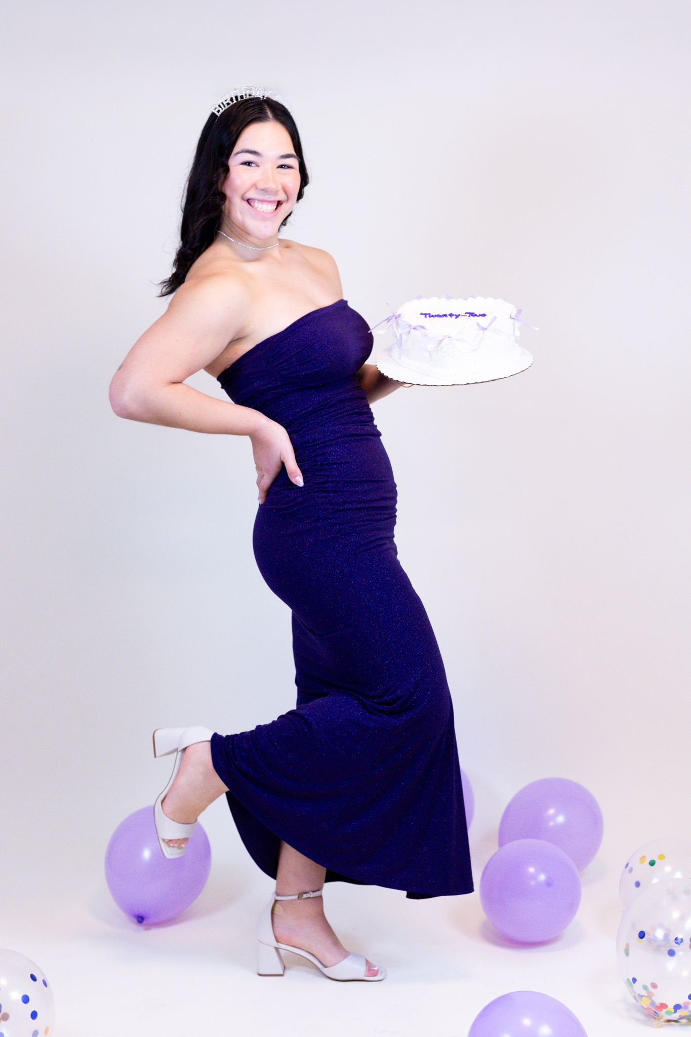 Maternity photoshoot featuring a woman in a navy dress posing with purple balloons and confetti against white backdrop.
