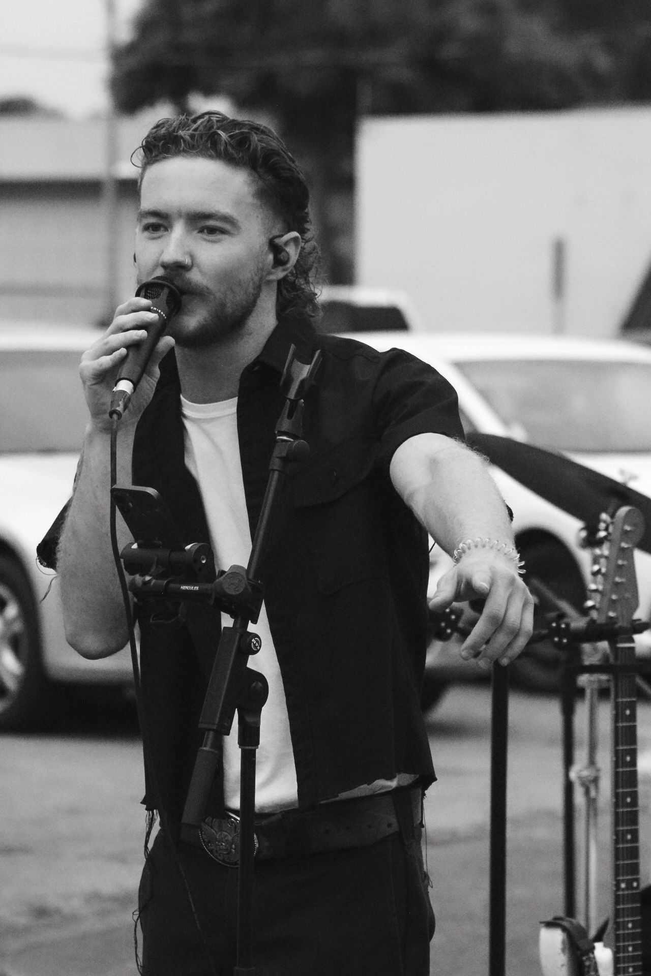 Black and white photo of a musician singing into a microphone while performing outdoors with guitars visible.