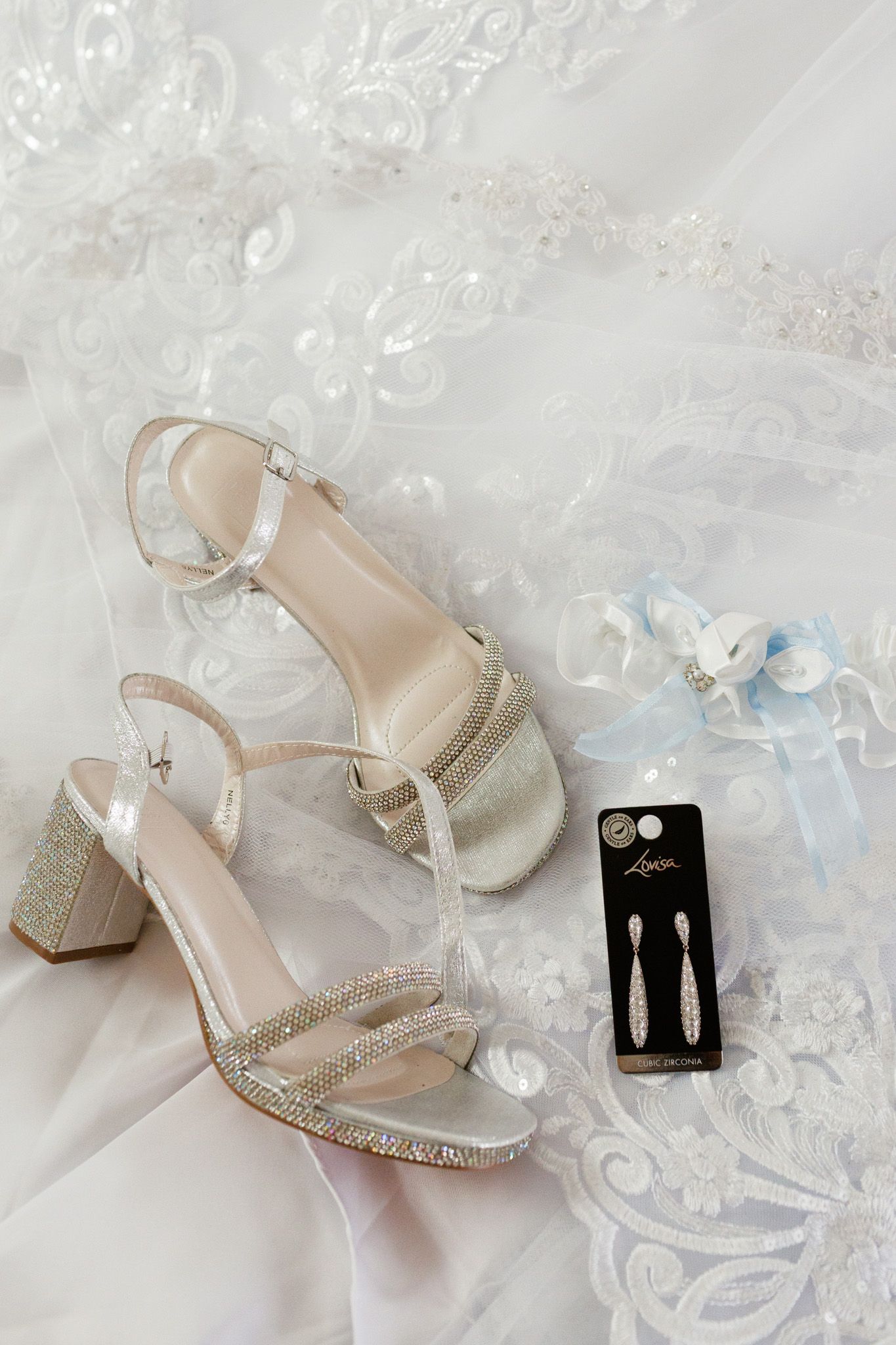 Wedding accessories including silver glitter sandals, dangle earrings, and a blue garter displayed on lace fabric.