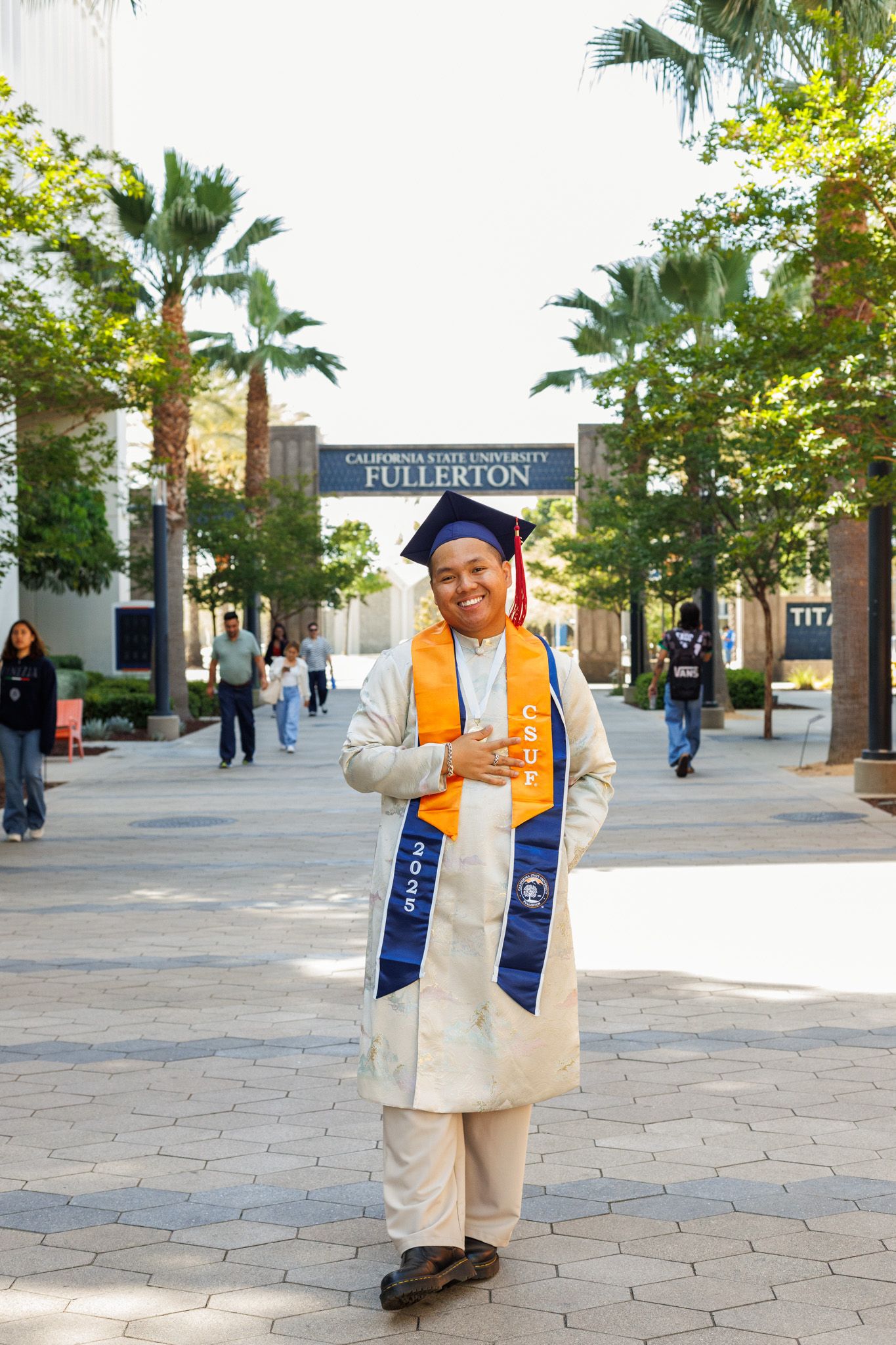 A graduate in cap and gown with graduation stoles stands on a brick pathway at Cal State Fullerton campus with palm trees.