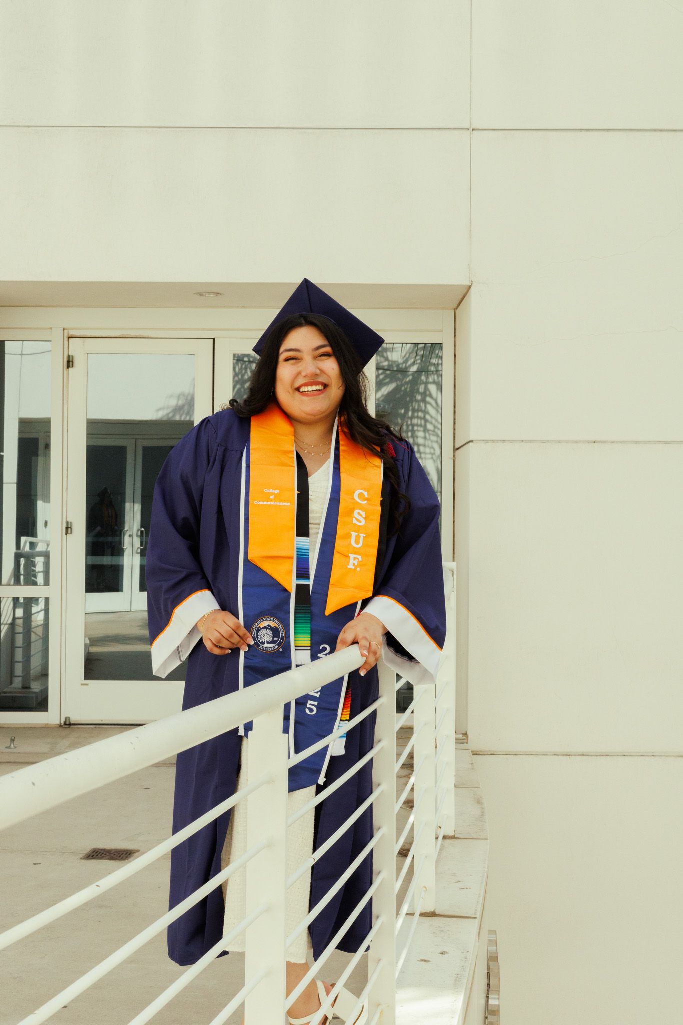 Graduate in navy blue cap and gown with gold stole stands smiling on outdoor balcony.