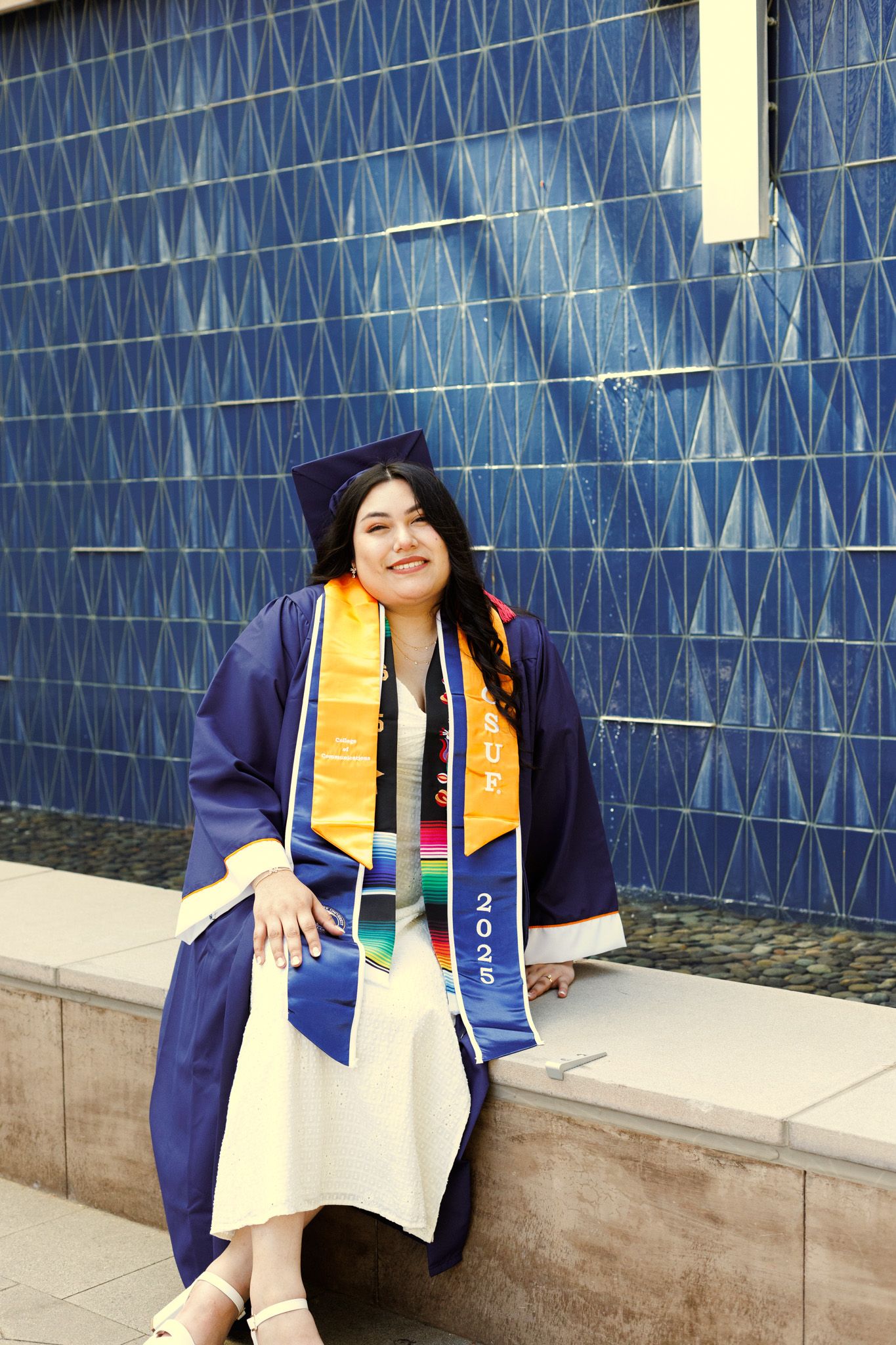 Graduate in blue cap and gown with gold stole poses in front of geometric blue wall during commencement ceremony.