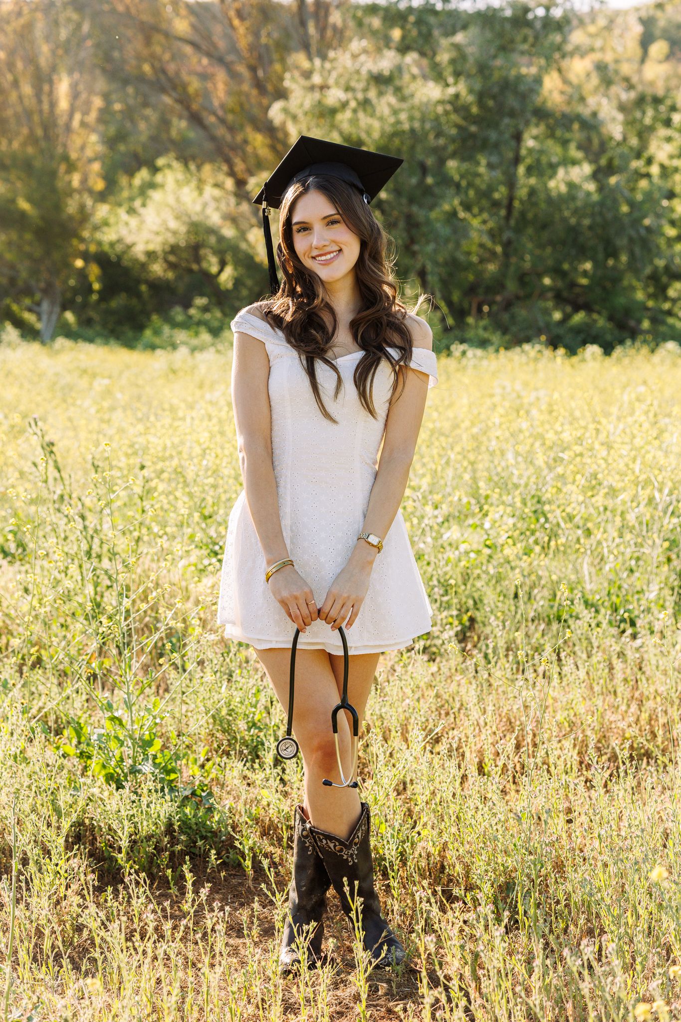Smiling graduate in white dress, mortarboard cap, cowboy boots, holding stethoscope in sunny field.
