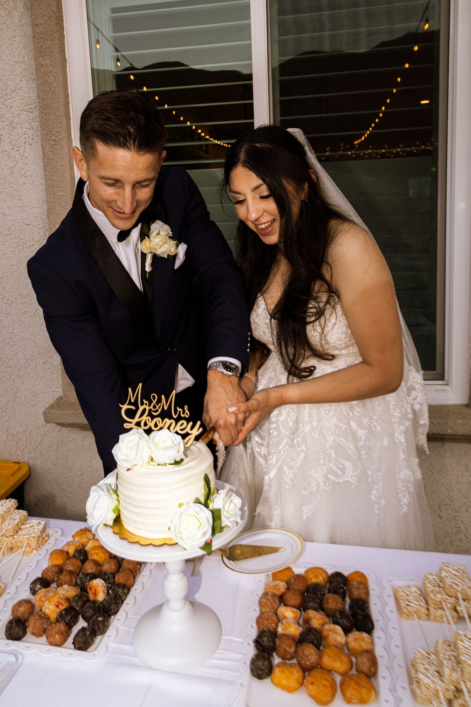 A couple in wedding attire cutting their white wedding cake together at their dessert table with festive lights in background.