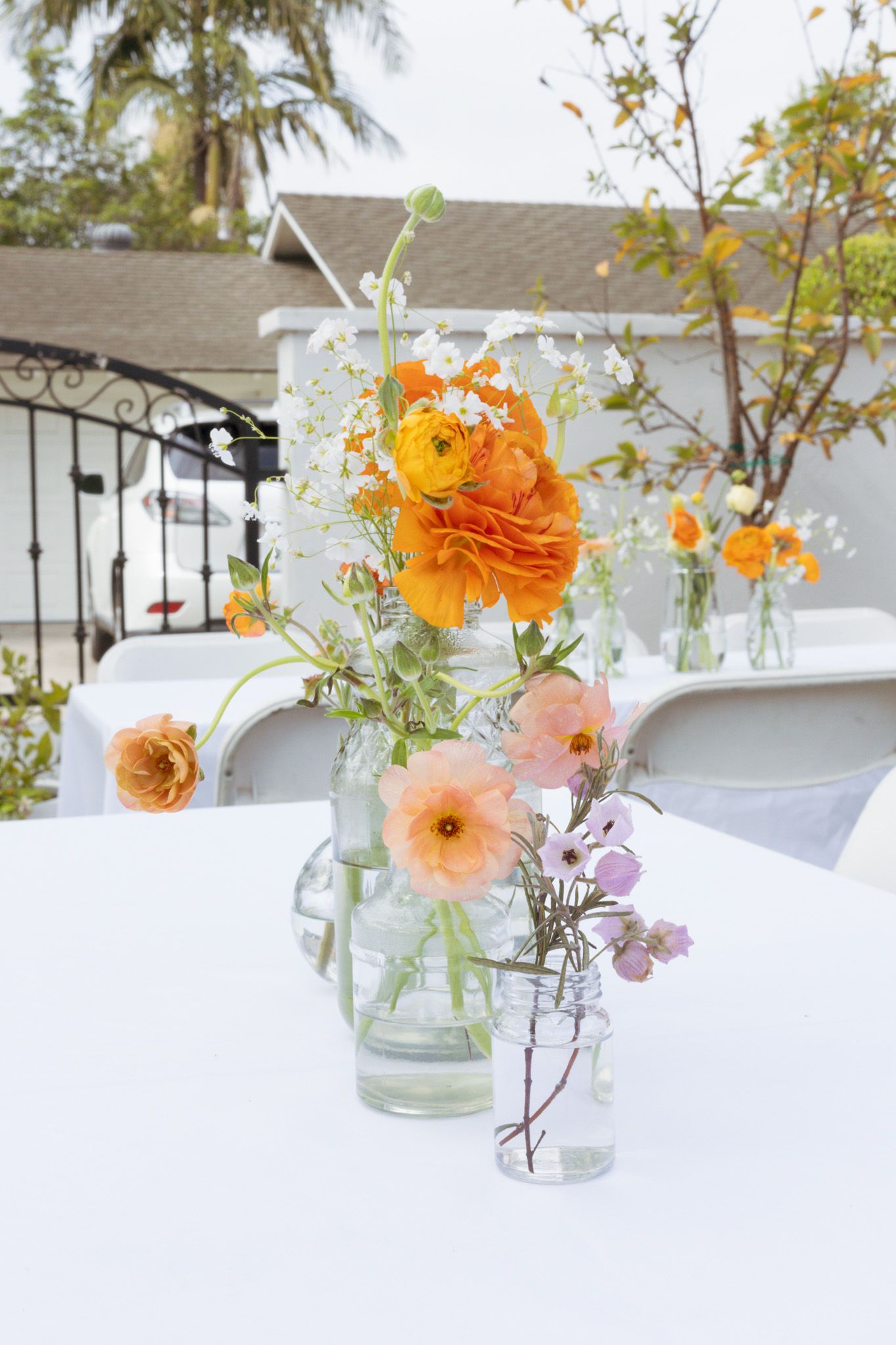 Glass vases with orange ranunculus and pink dahlia flowers arranged on a white table outdoors with a house in background.