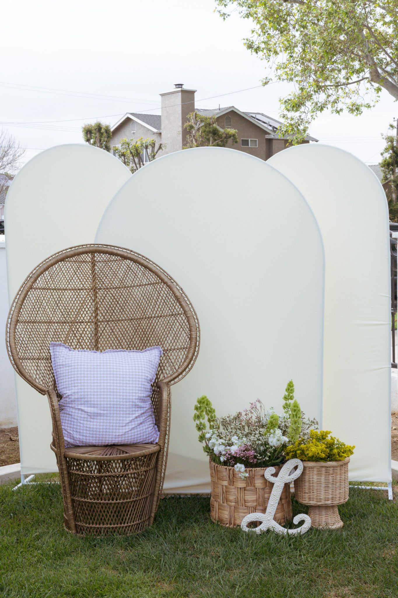 Vintage wicker peacock chair with purple pillow sits against white arch backdrop with potted plants in garden setting.