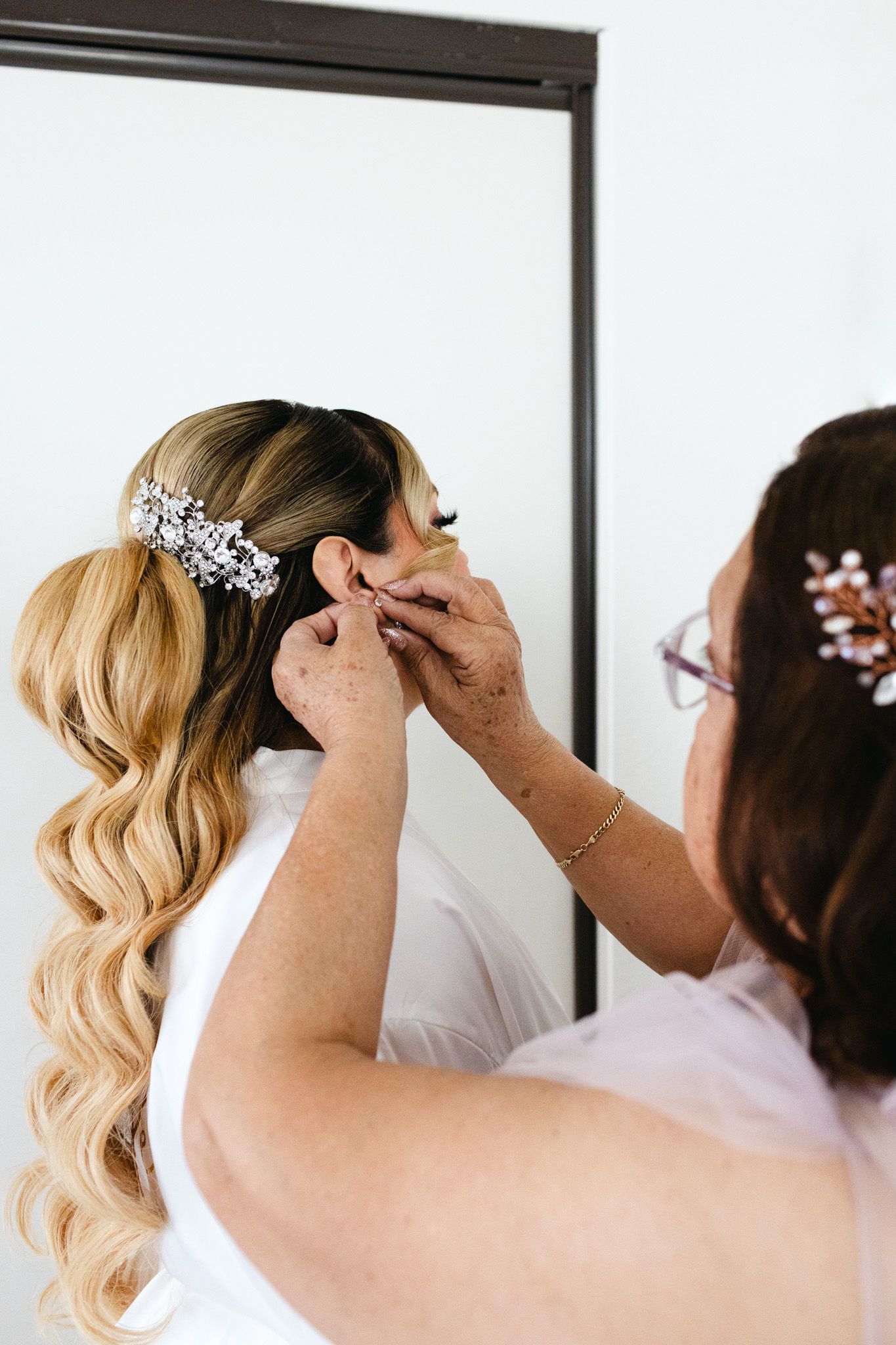 A stylist adjusts a crystal floral hair accessory on long blonde wavy hair during bridal preparation.