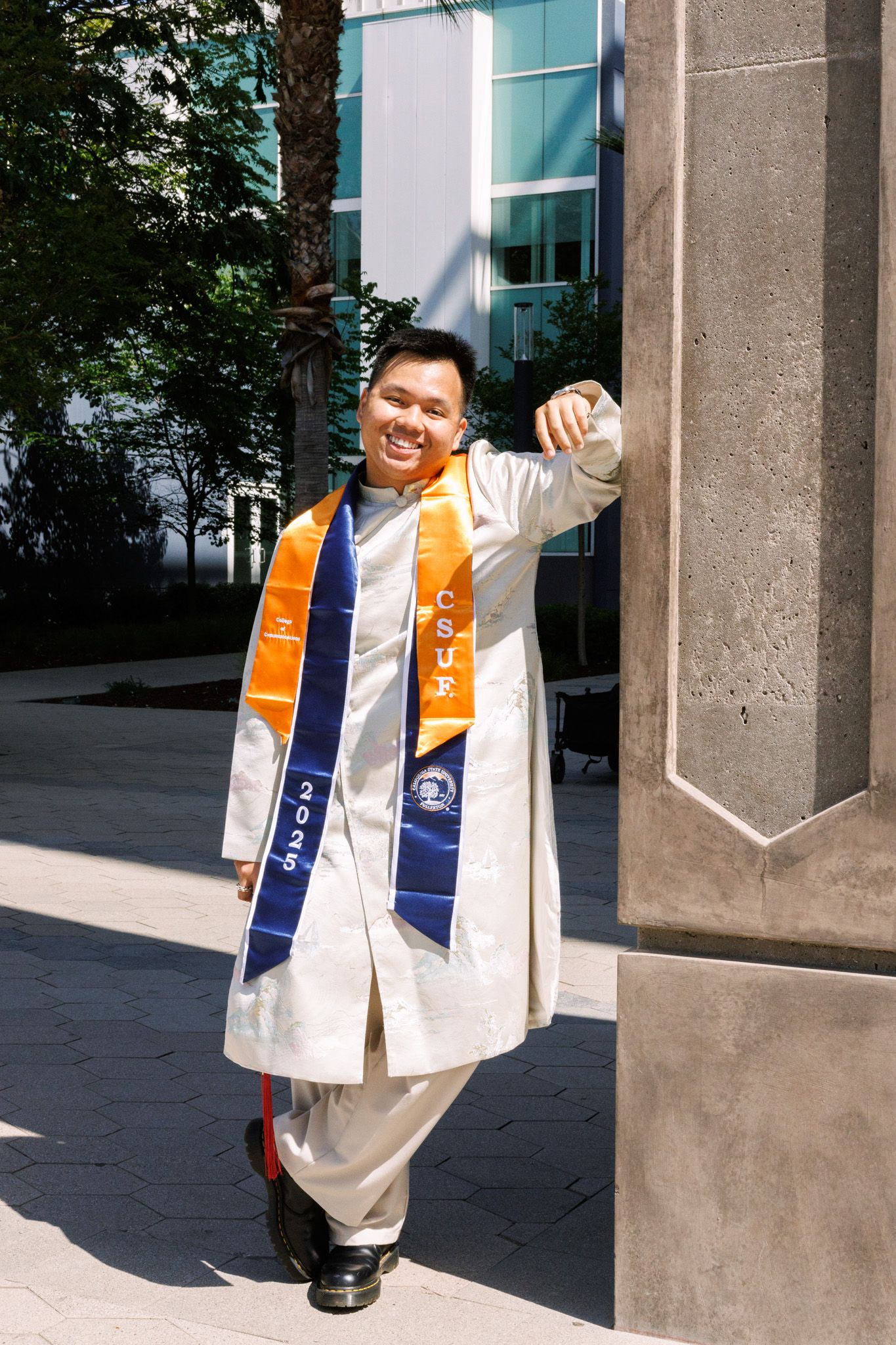 Graduate wearing white academic regalia with blue and orange stolas poses joyfully outside a modern building.
