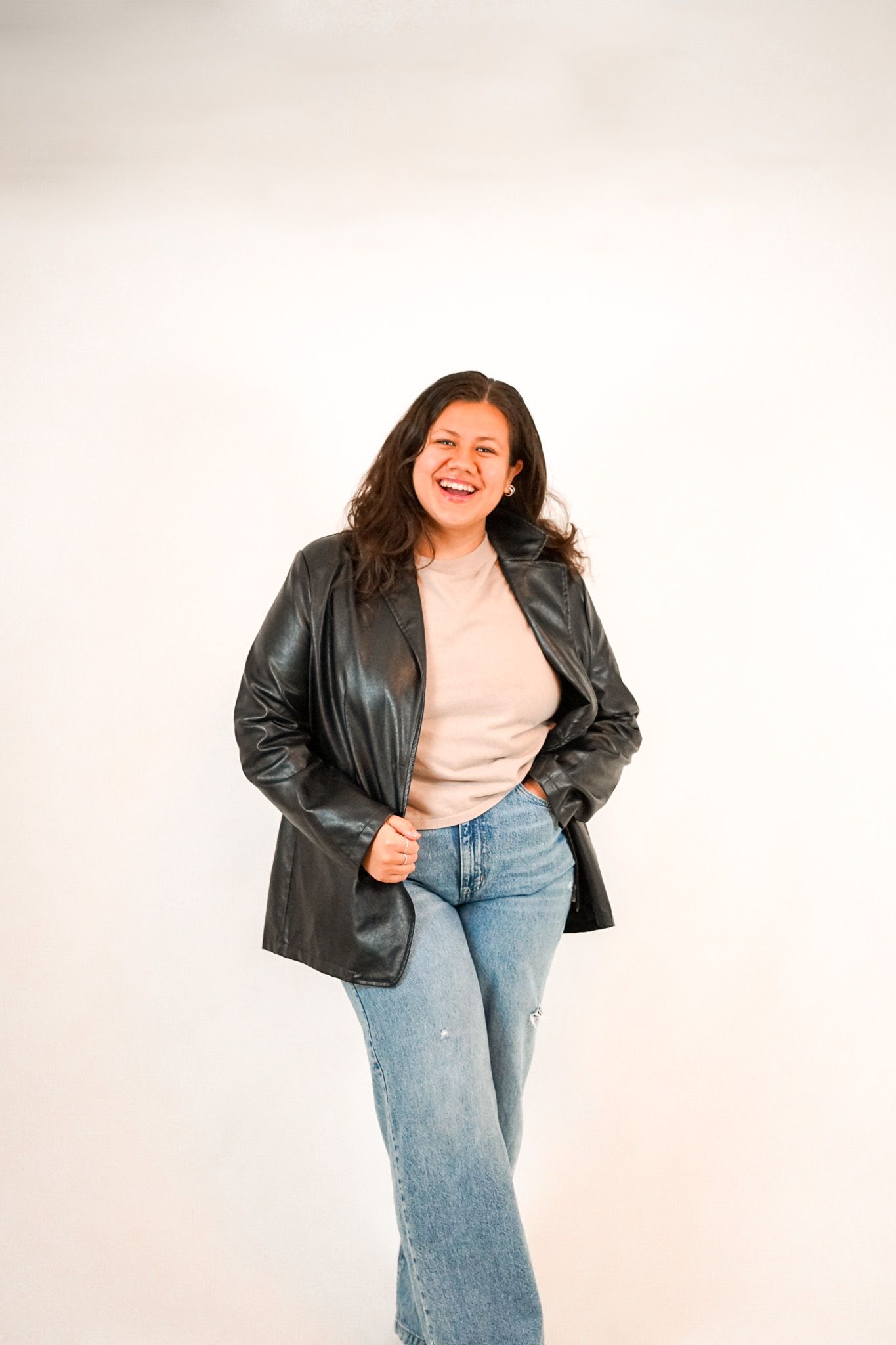A person in a black leather jacket and jeans poses casually against a white backdrop with a cheerful expression.