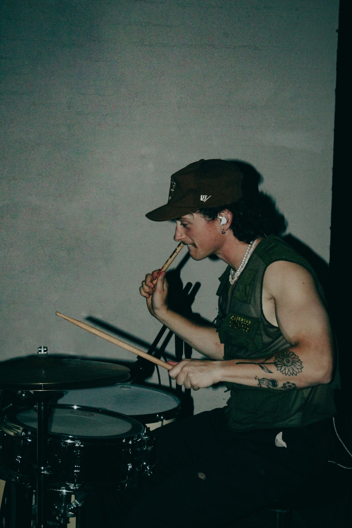 Drummer in a baseball cap and tank top plays drums in dimly lit setting.