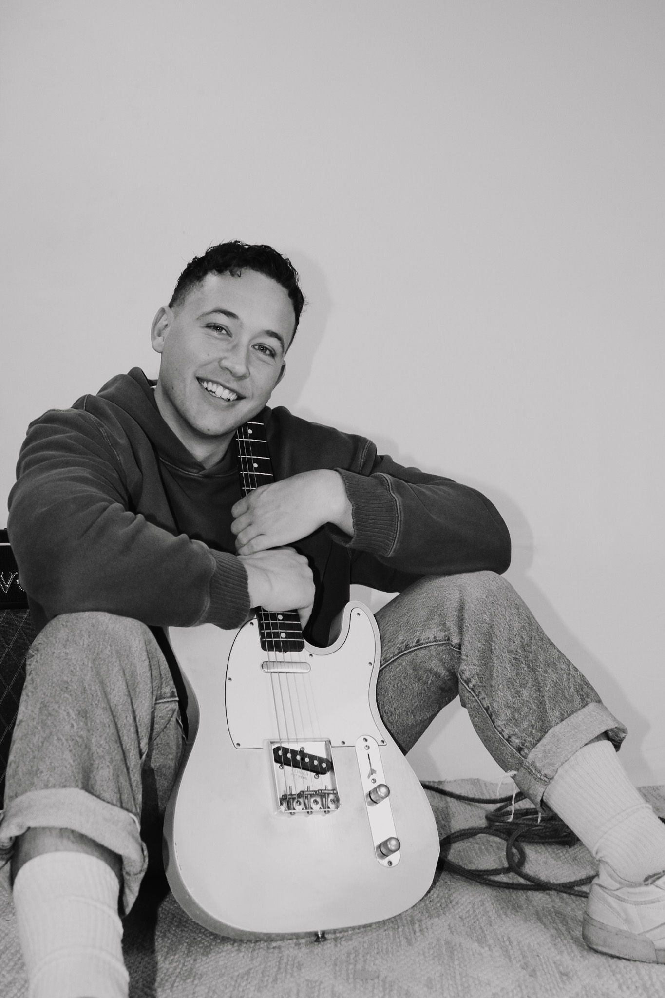 A black and white portrait of someone sitting cross-legged with a white electric guitar.