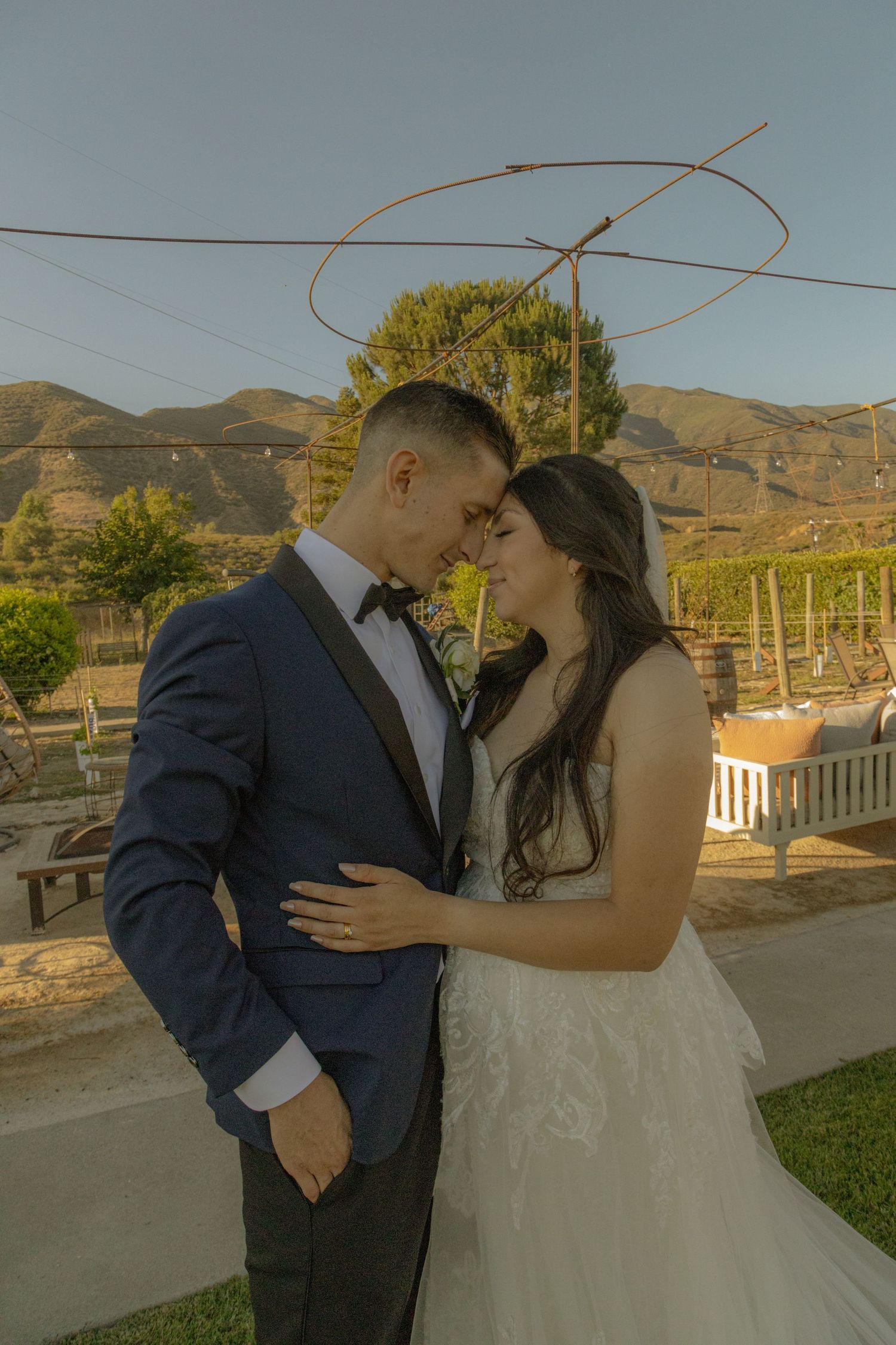 Two people in wedding attire kiss in front of a scenic mountain landscape at sunset on an outdoor deck.