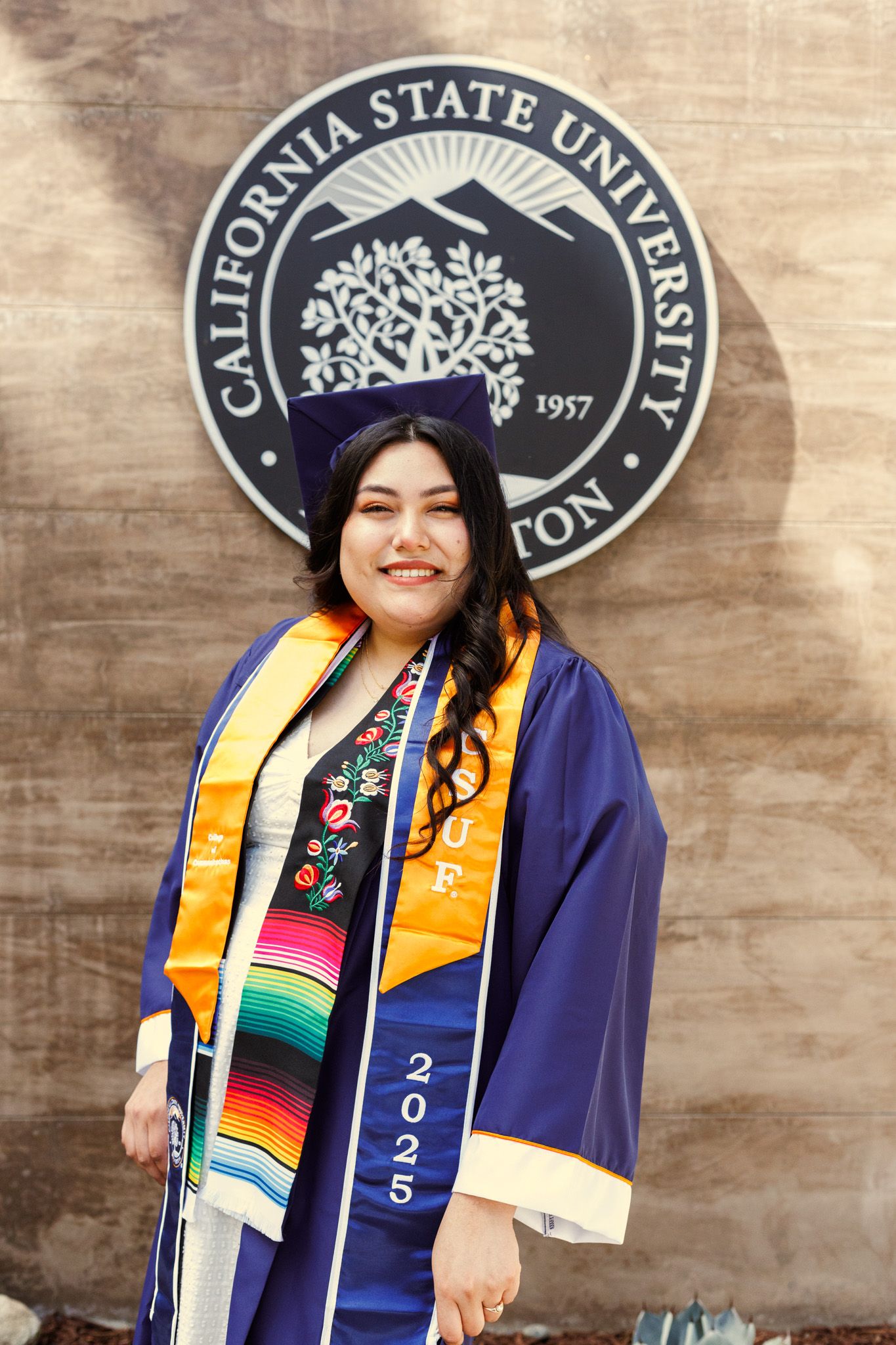 Graduate in blue cap and gown with colorful stoles poses in front of California State University seal at graduation ceremony.