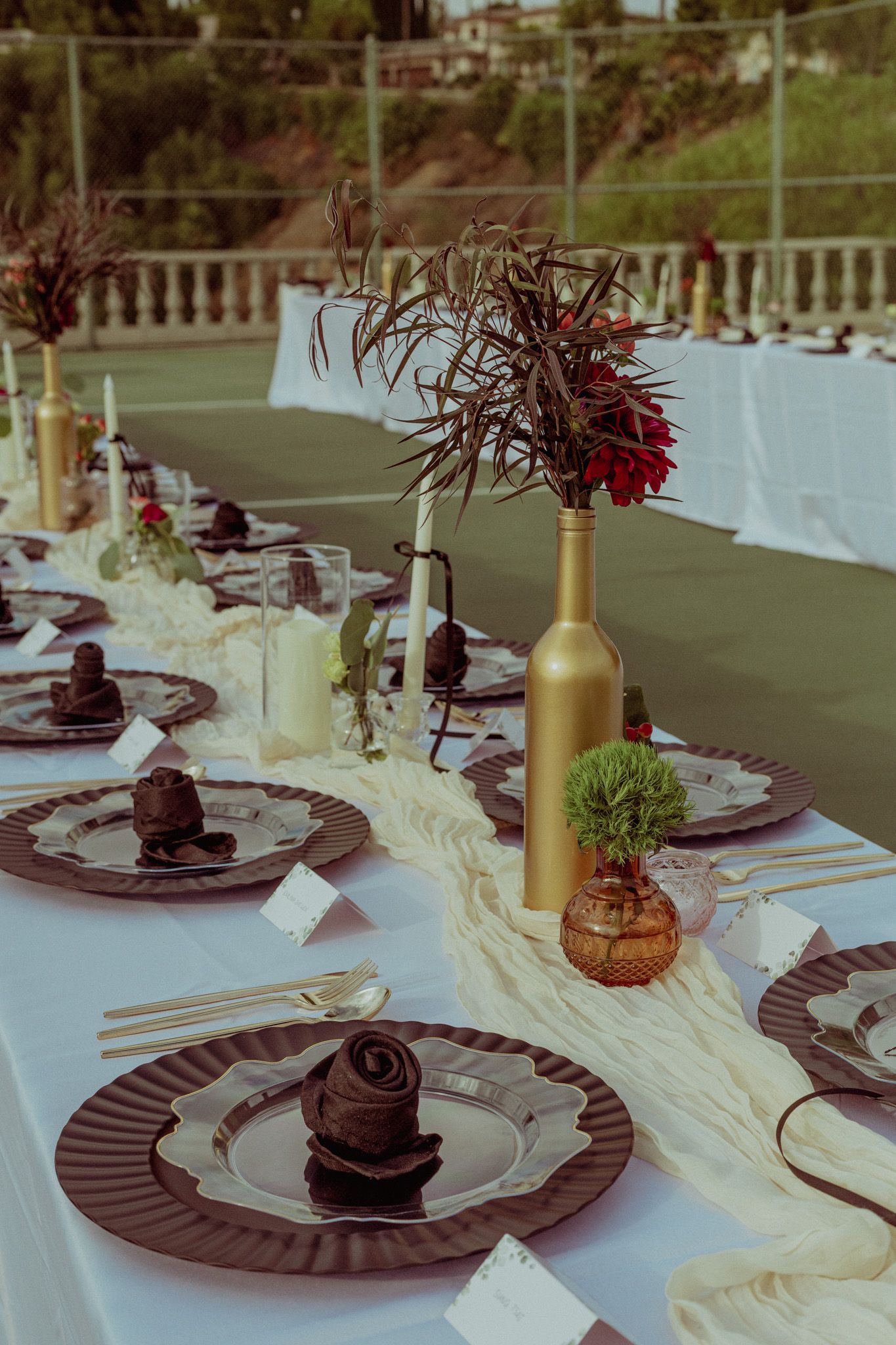 Elegant wedding reception table settings with dark charger plates and gold bottles on cream tablecloth runners.