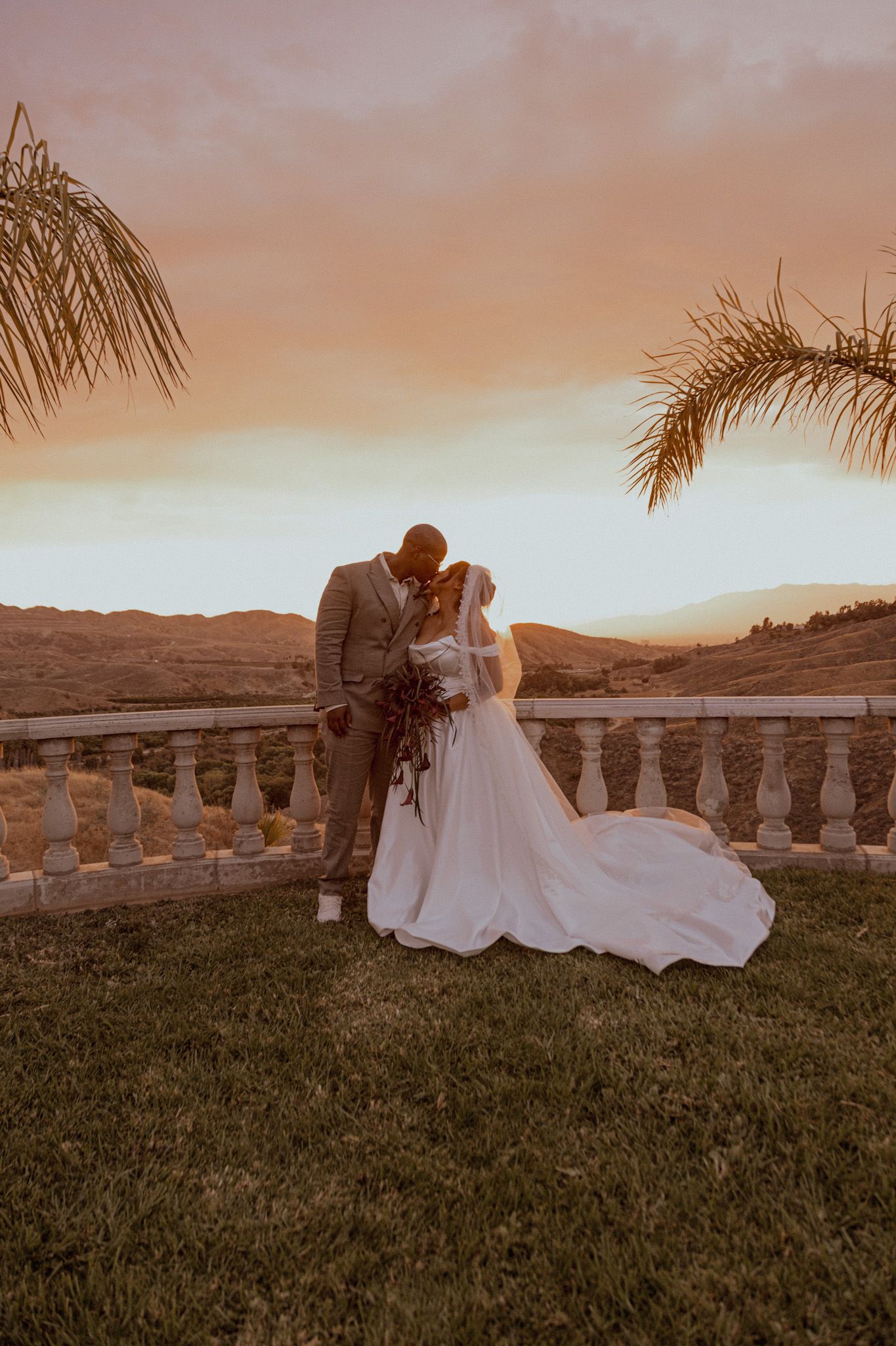 A couple shares a romantic moment at sunset overlooking a scenic vista with palm trees and mountains in the background.