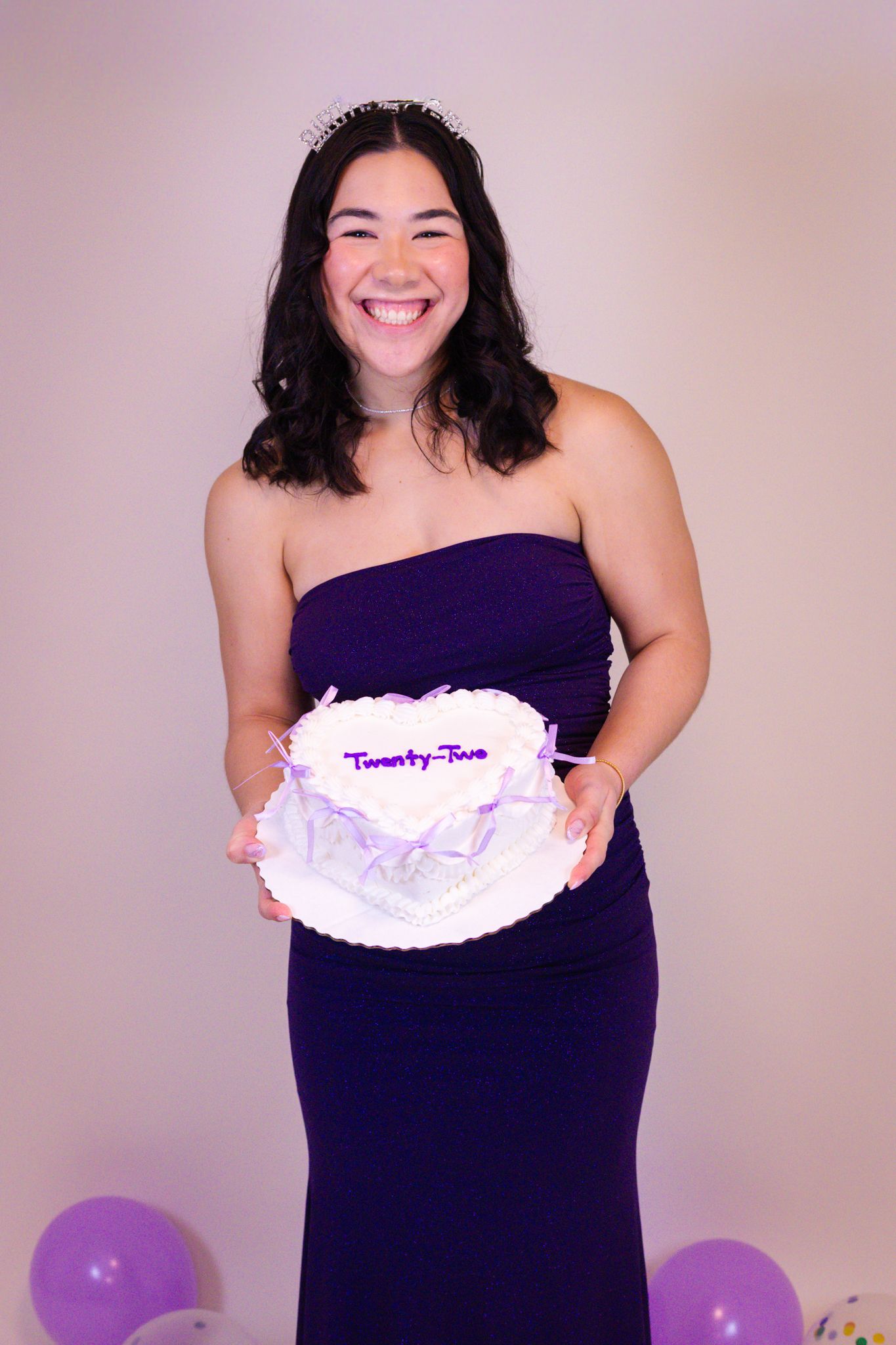 A person in a dark purple dress holds a white cake while posing in front of a light colored wall.
