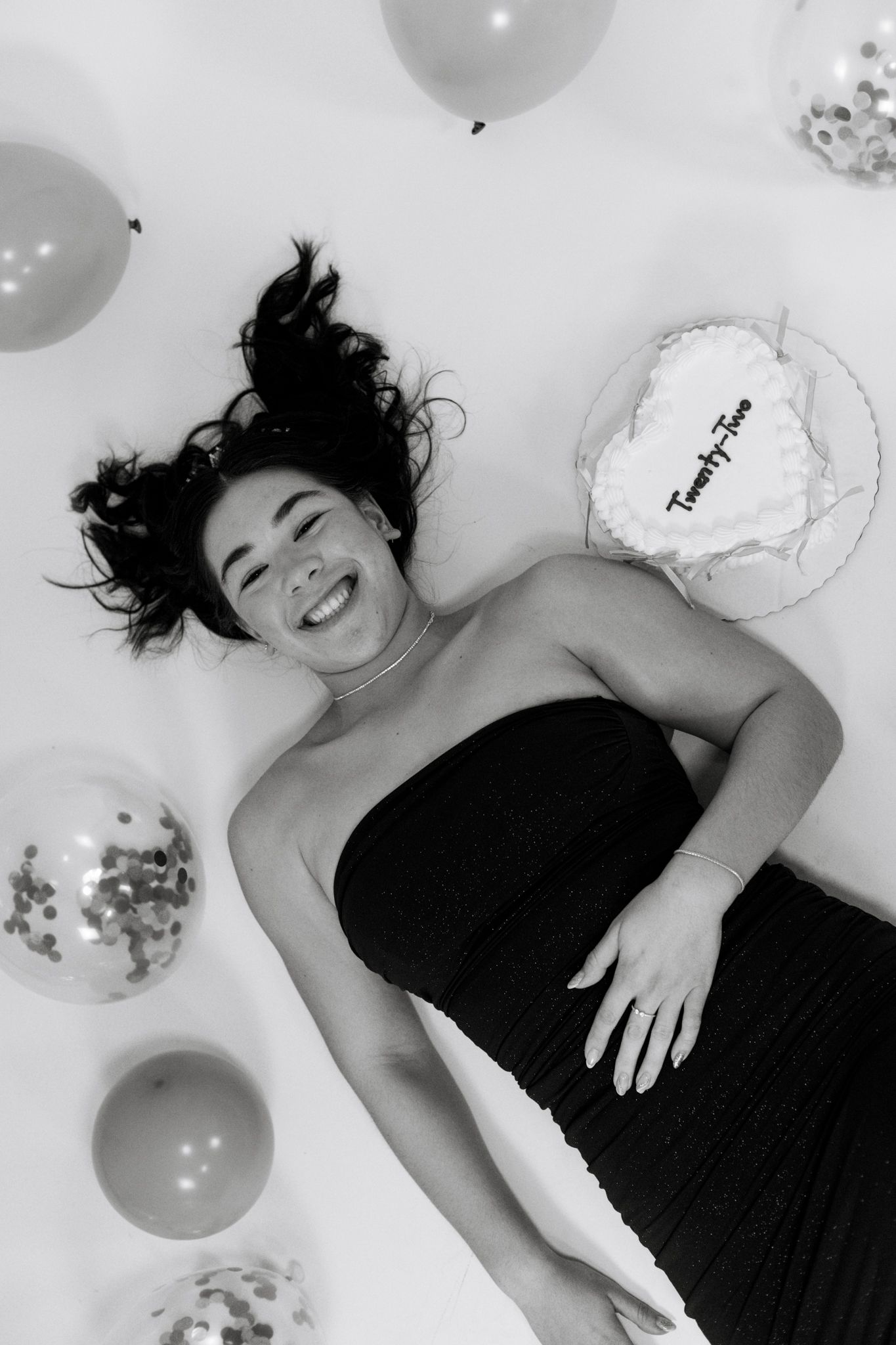 Black and white photo of someone lying down surrounded by balloons and confetti in a celebratory setting.