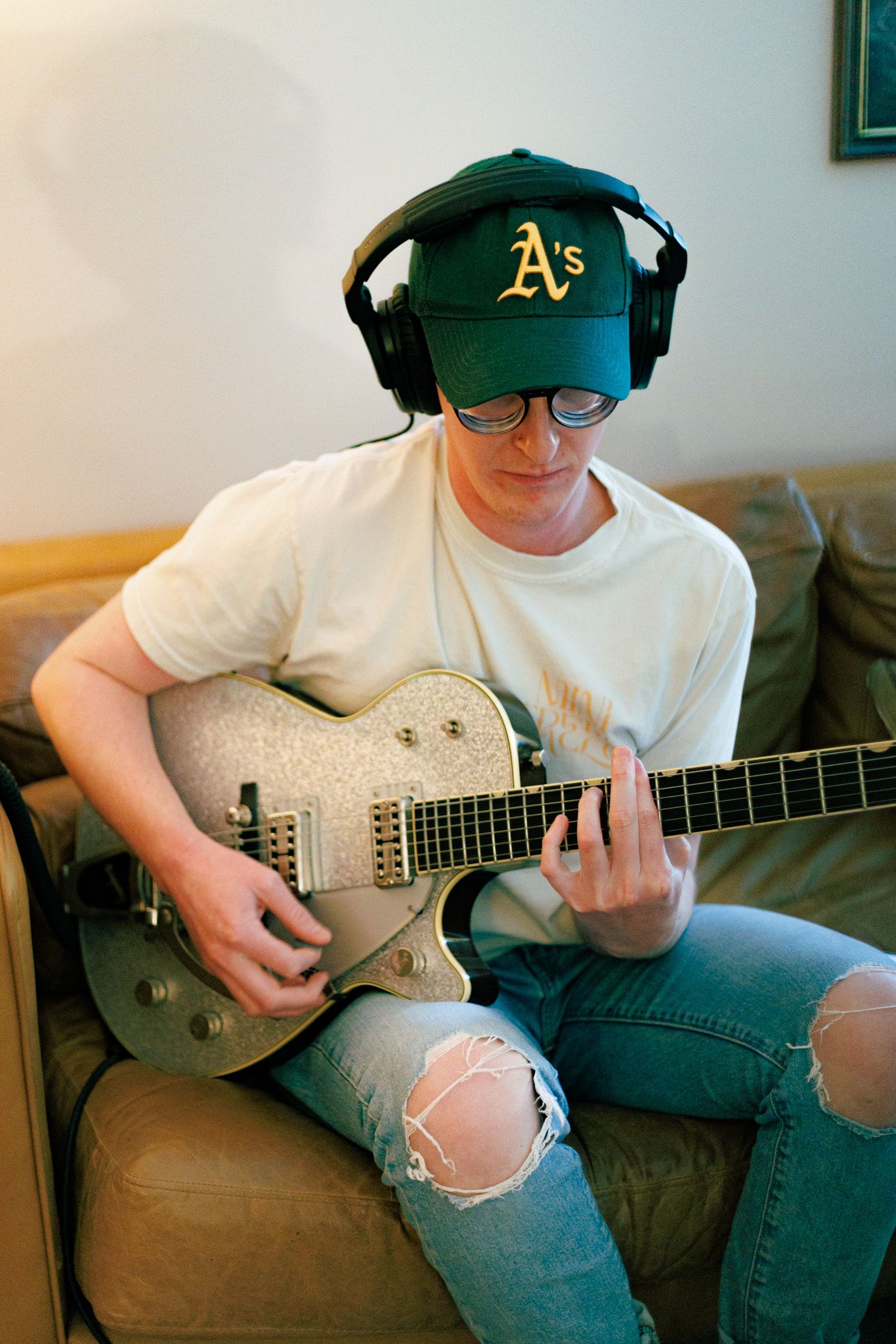 Guitar player wearing Oakland A's cap and headphones plays silver electric guitar while sitting on gray couch in casual attire.