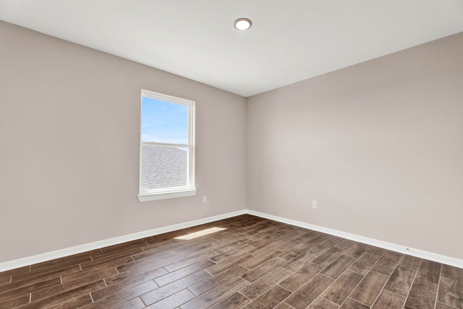 Empty room with wood-look tile flooring, neutral beige walls, white baseboards, and a single window allowing natural light.