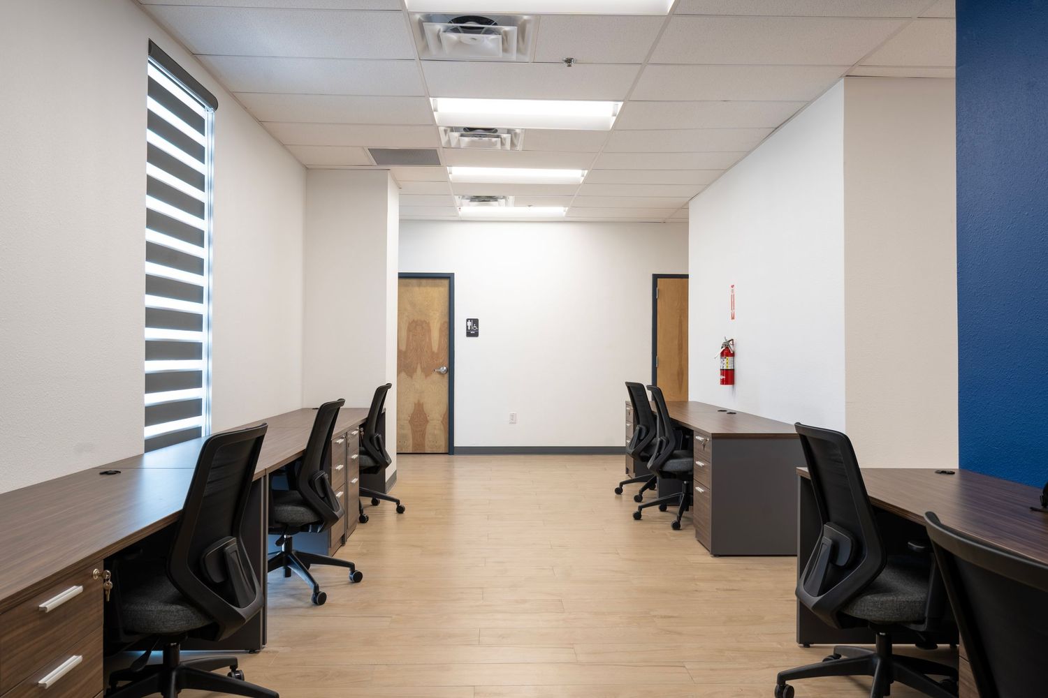 Empty office workspace with black rolling chairs and wooden desks along a white hallway with fluorescent lighting.