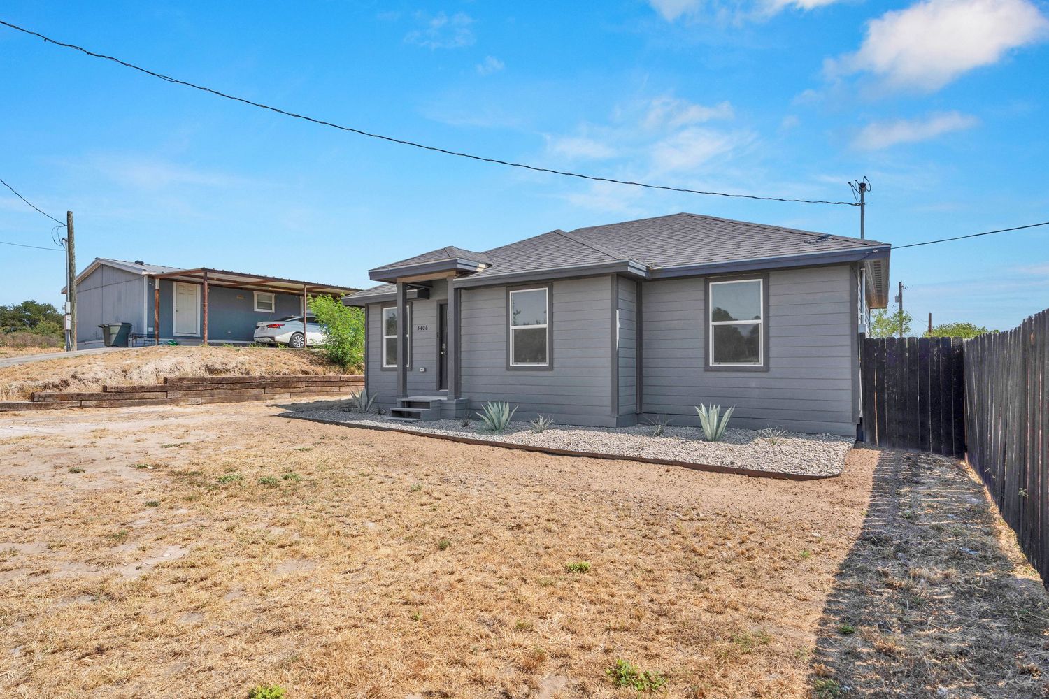 Gray ranch-style home with a gravel yard, wood fence, and blue sky in a residential neighborhood.