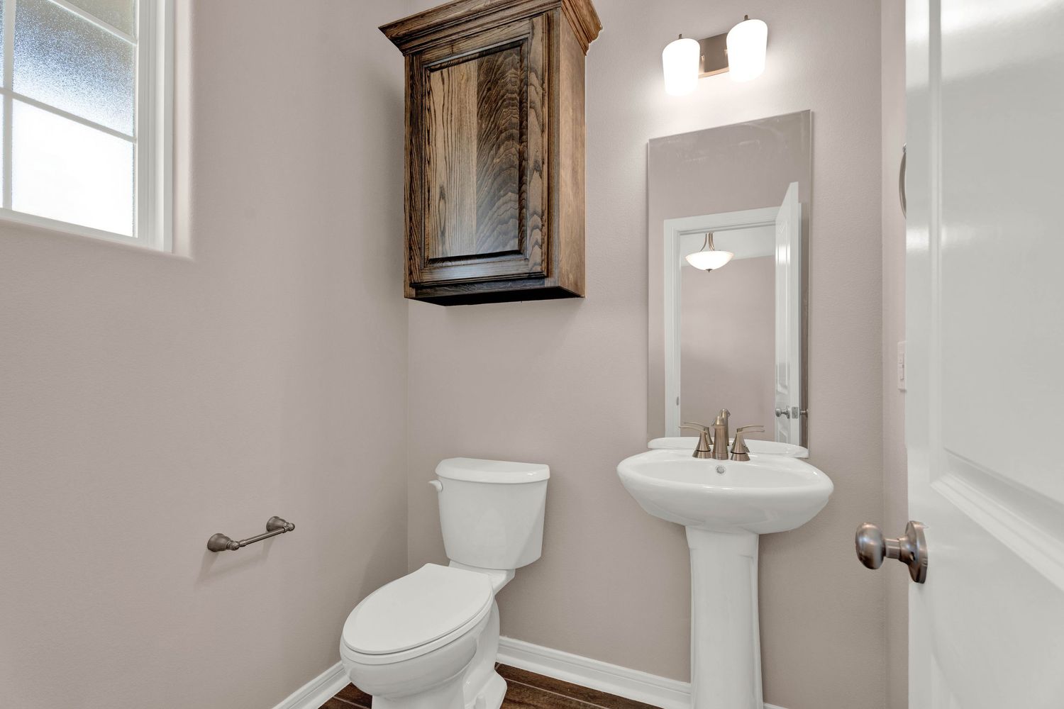 Small bathroom with pedestal sink, white toilet, rustic wood wall cabinet, and framed mirror on beige walls.