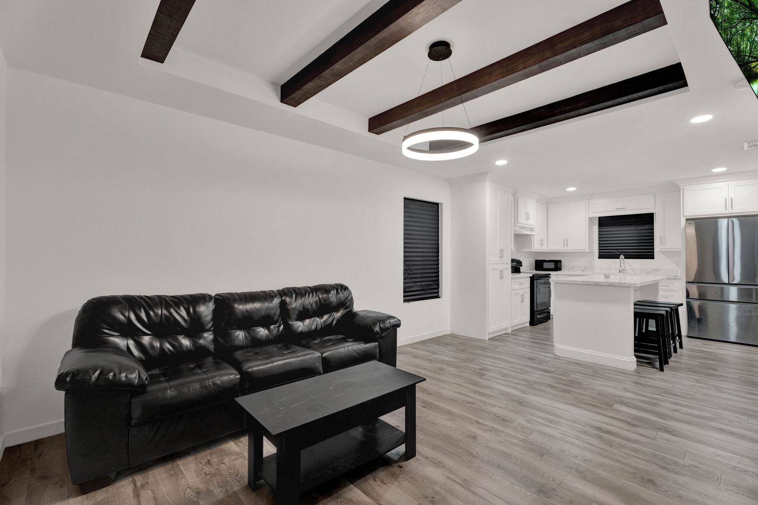 Modern living room with black leather sofa, wood beam ceiling, and open-concept kitchen with white cabinets and stainless appliances.
