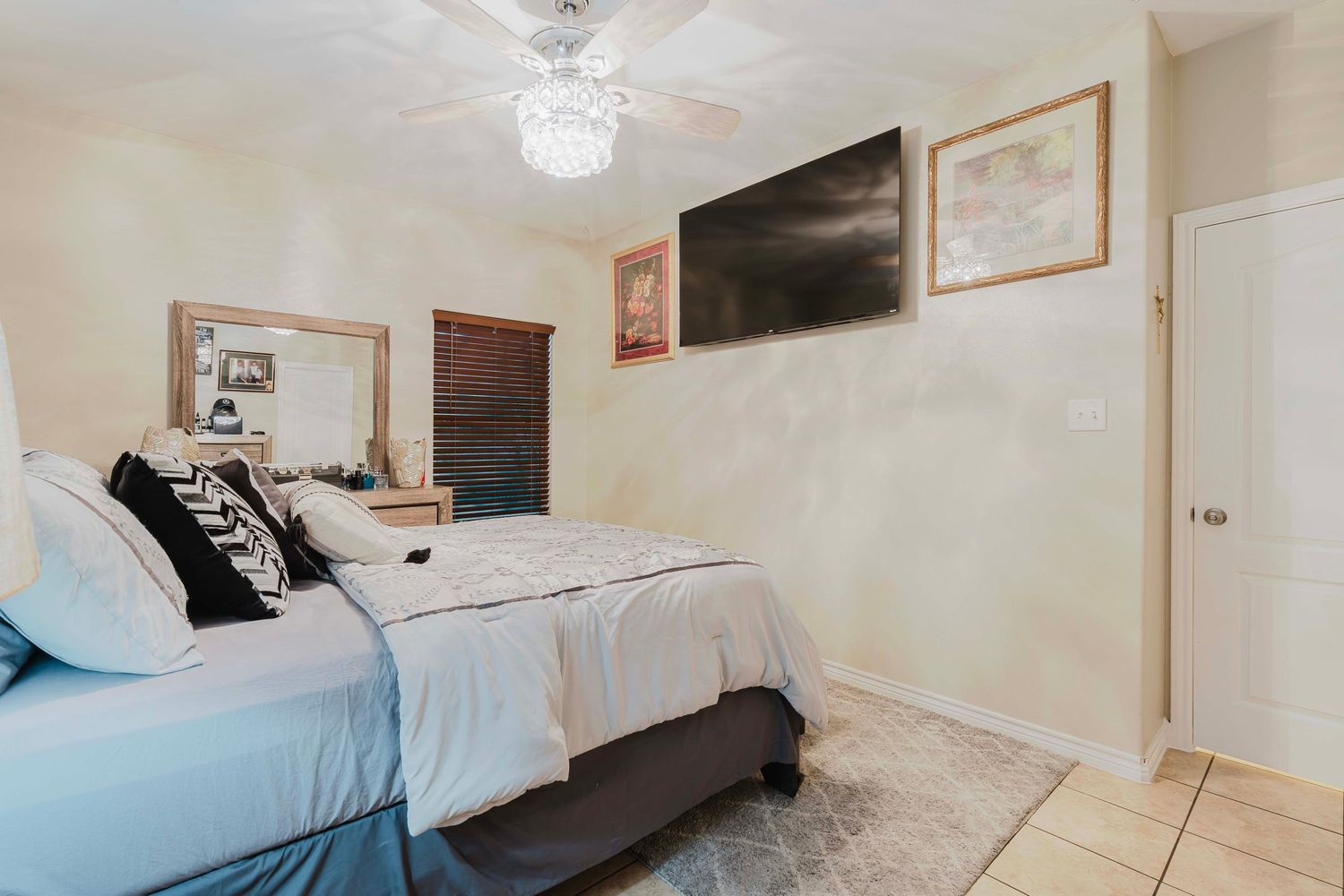 Modern bedroom interior with mounted TV, crystal chandelier, and light blue bedding in a tiled floor space.