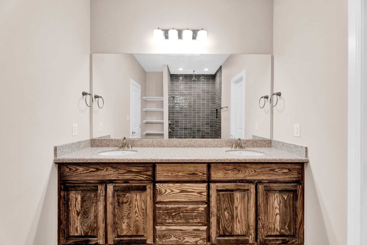 Double sink bathroom vanity with dark wood cabinets, granite countertop, large mirror, and tiled walk-in shower.