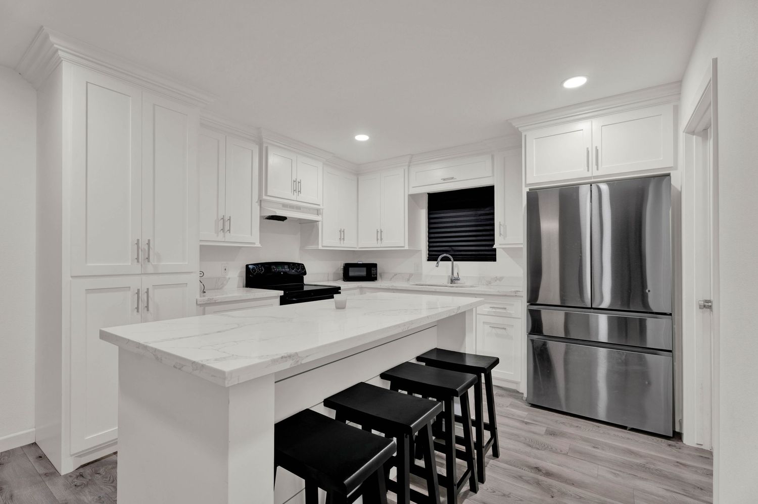 Modern white kitchen with marble island, black bar stools, stainless steel fridge, and white shaker cabinets on hardwood floors.
