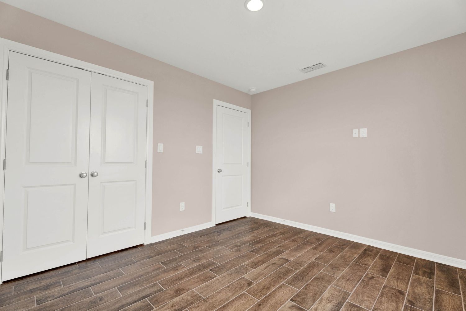 Empty bedroom with wood-look tile floors, white bifold closet doors, and soft pink walls in a modern home.