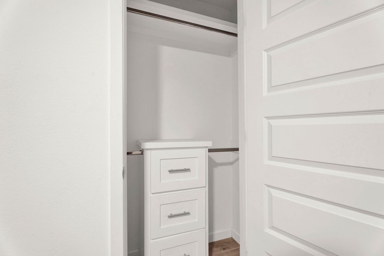 Small walk-in closet with white walls, hanging rod, and a white three-drawer dresser.