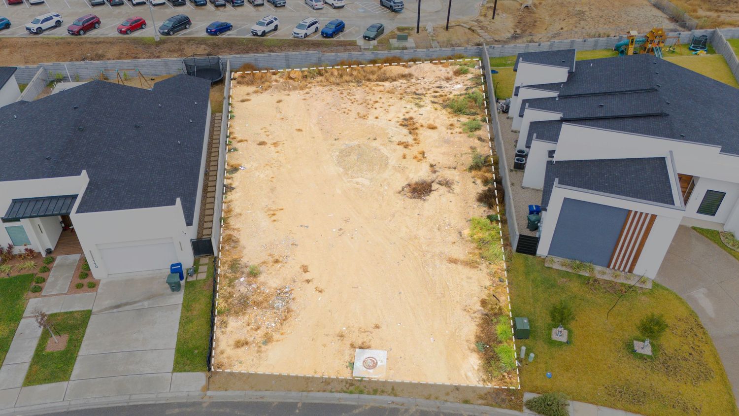 Aerial view of a vacant sandy lot between modern residential houses with gray roofs and driveways.