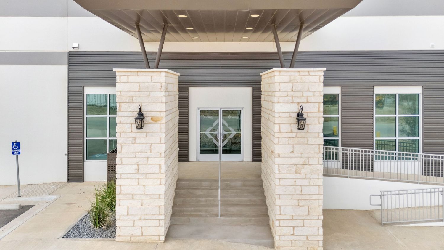 Modern building entrance with limestone pillars, glass doors, and wall sconces flanking a covered entryway.