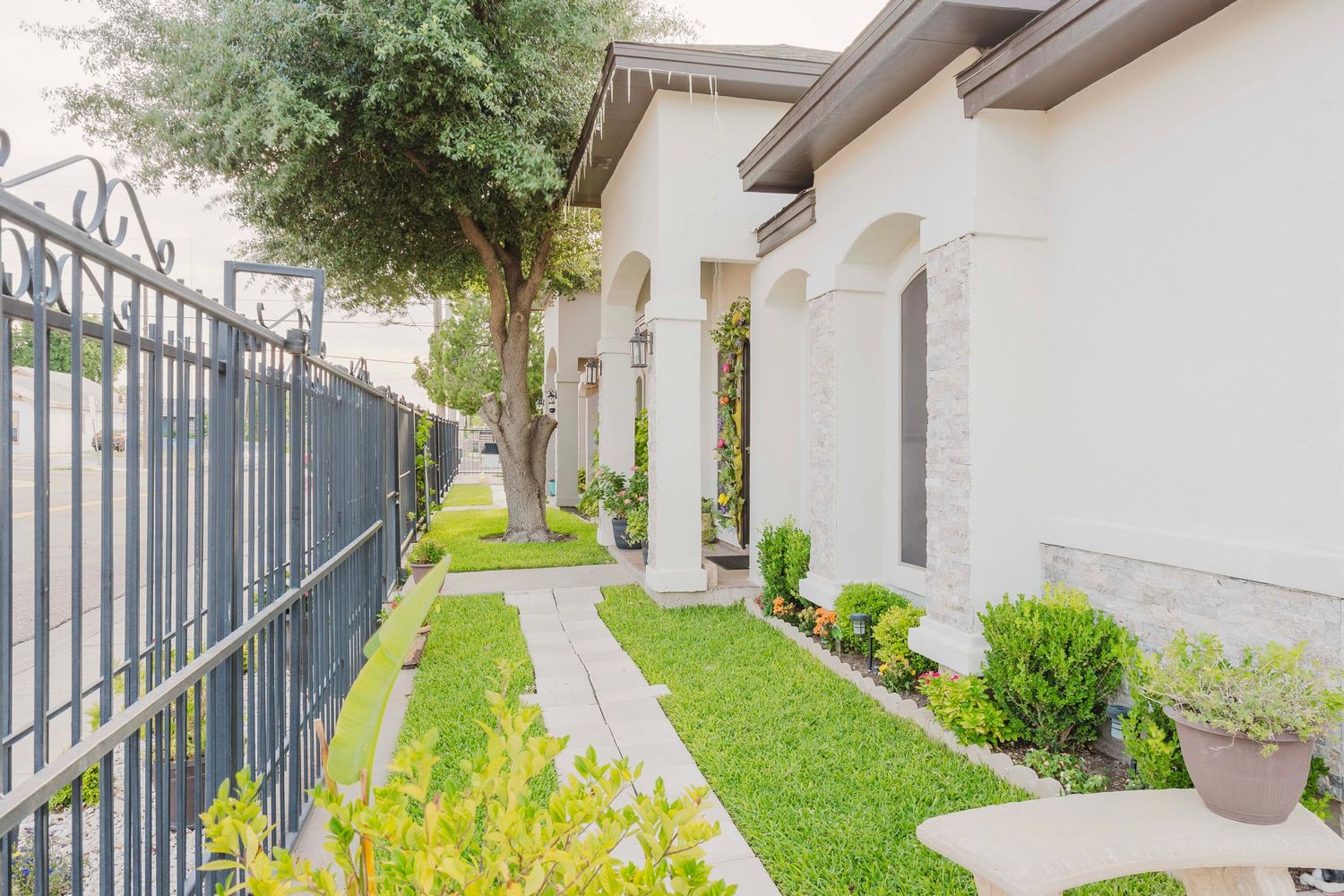 A well-maintained pathway with green lawn and decorative shrubs runs alongside a white house with wrought iron fencing.