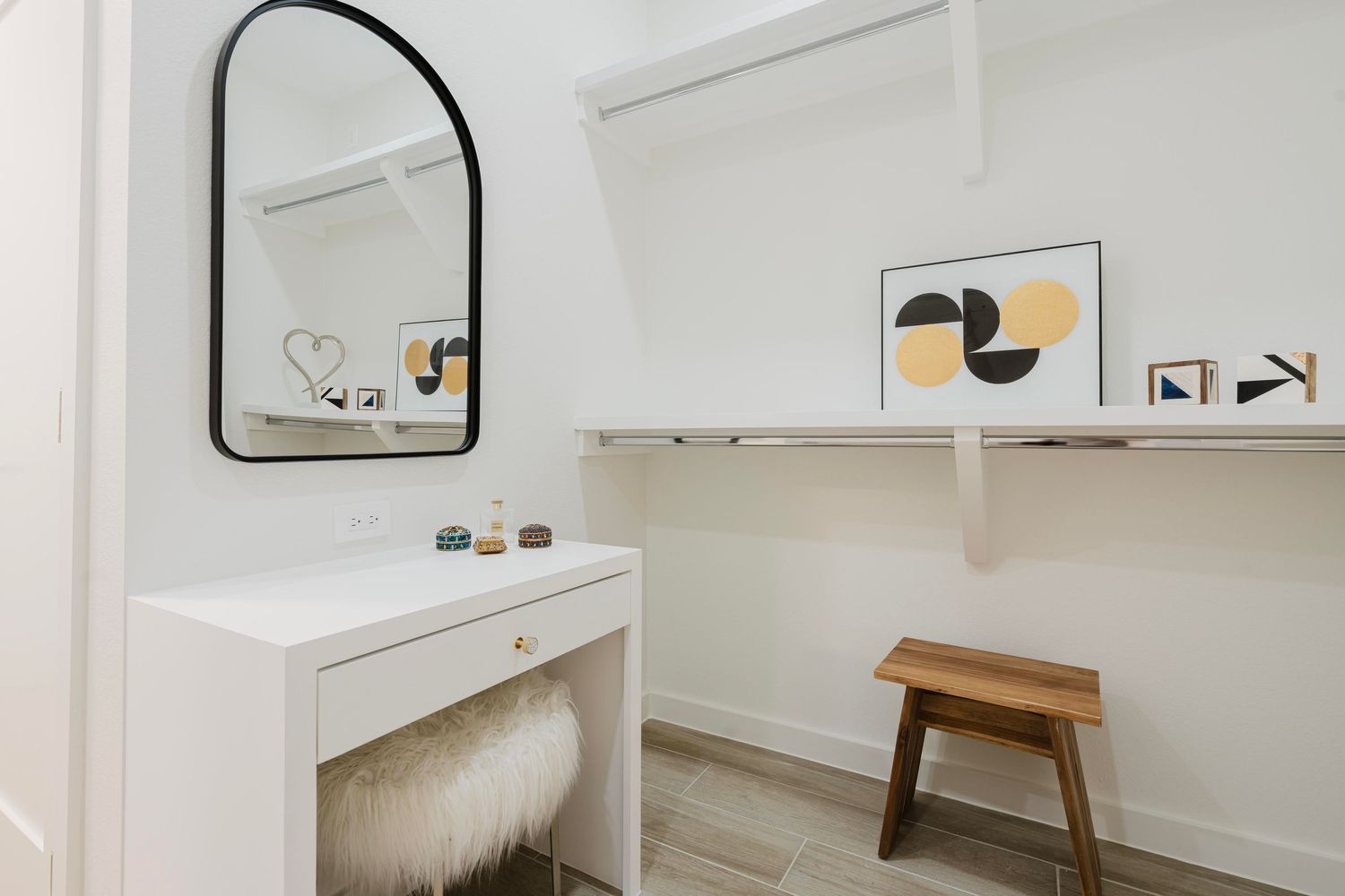 Elegant vanity space with furry stool, black arched mirror, and floating shelves displaying artwork.