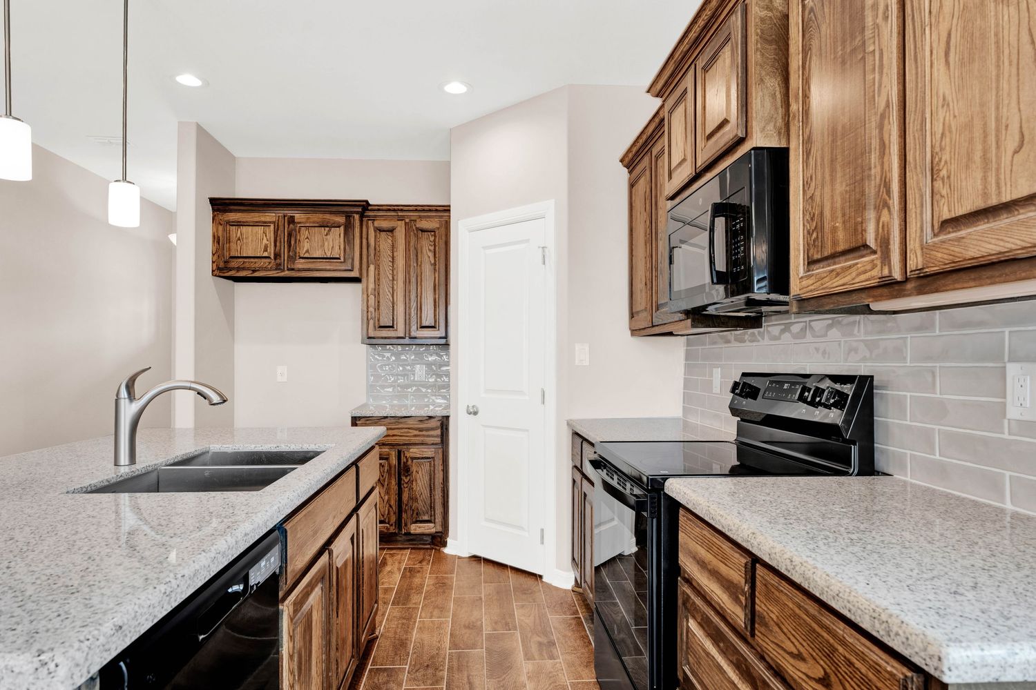 Modern kitchen with brown wood cabinets, granite countertops, black appliances, and subway tile backsplash on hardwood floors.