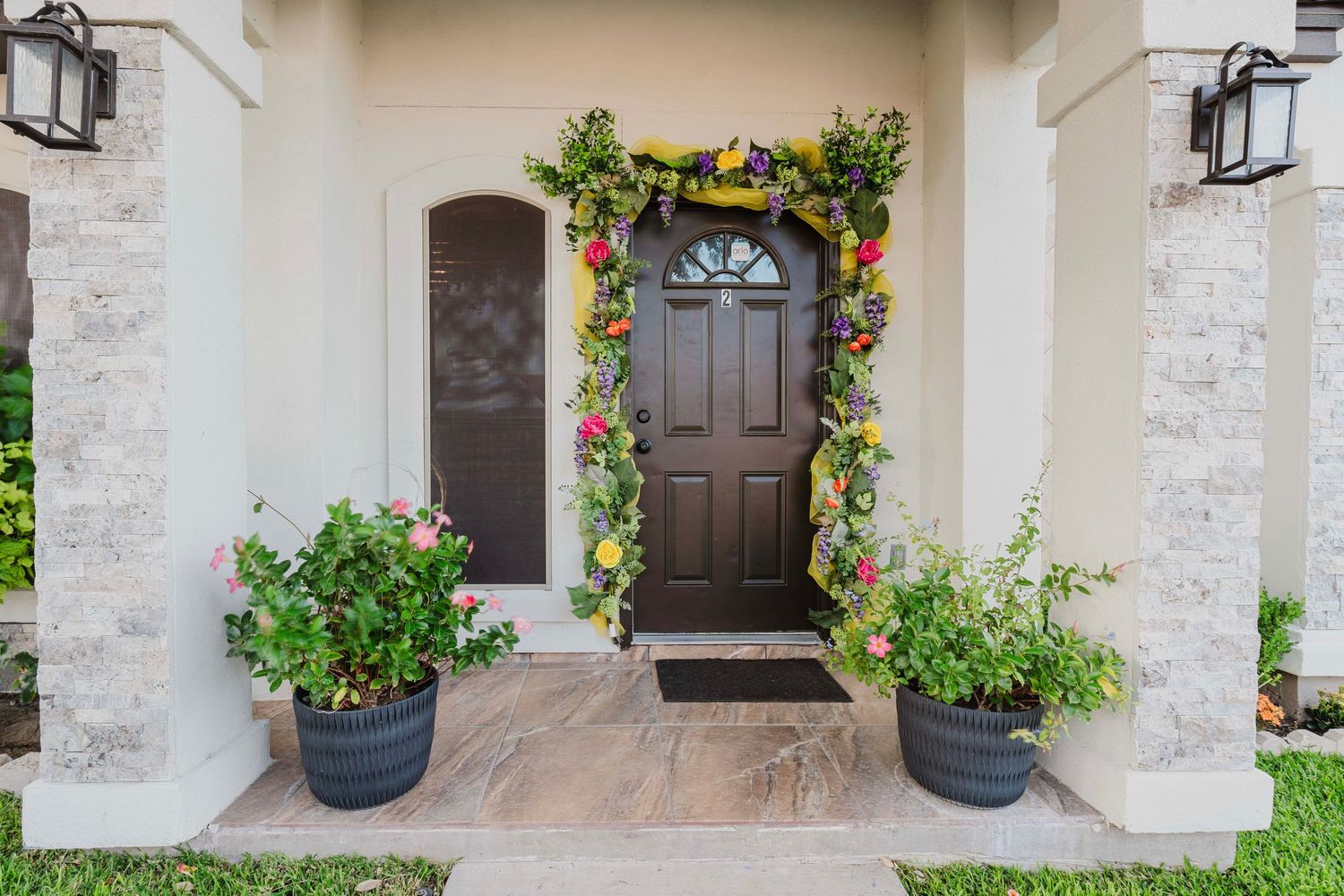 A dark brown front door framed by colorful floral garland and potted plants on a stone porch entrance.